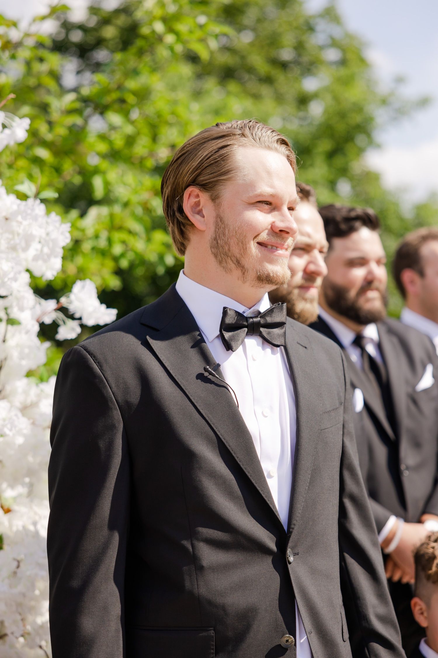Groomsmen in classic black tuxedos stand at an elegant outdoor wedding ceremony with white floral decorations.