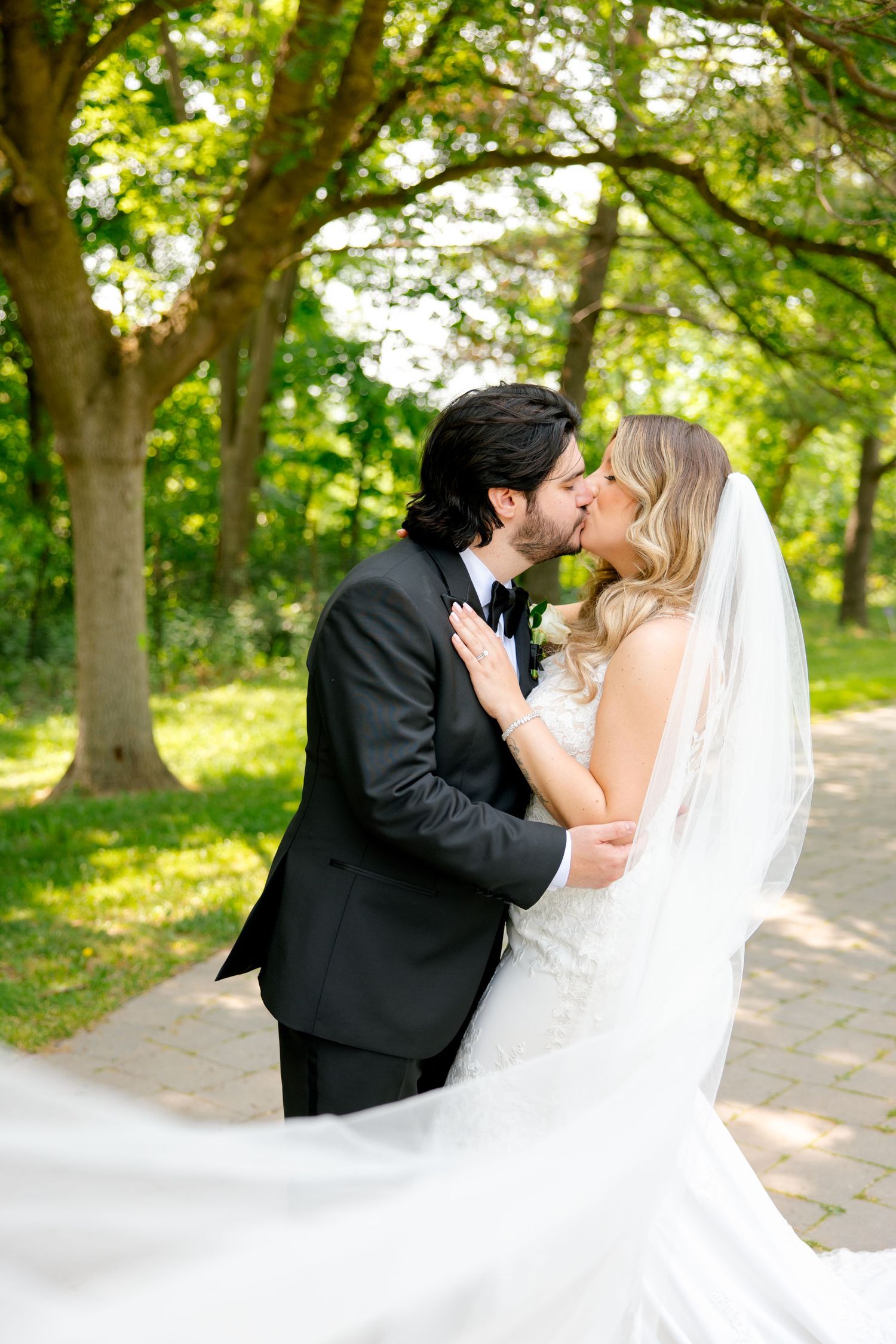 A romantic wedding kiss between newlyweds in a sun-dappled garden setting.