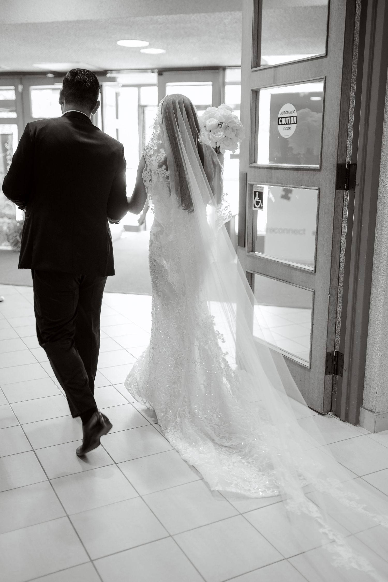 A black and white photo of newlyweds walking through a hallway with the bride's veil trailing behind.