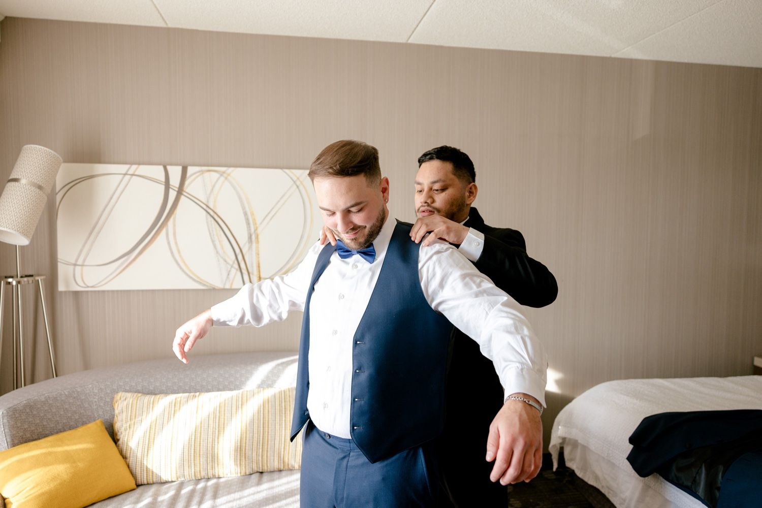 Two groomsmen share a playful moment in formal attire in a hotel room.