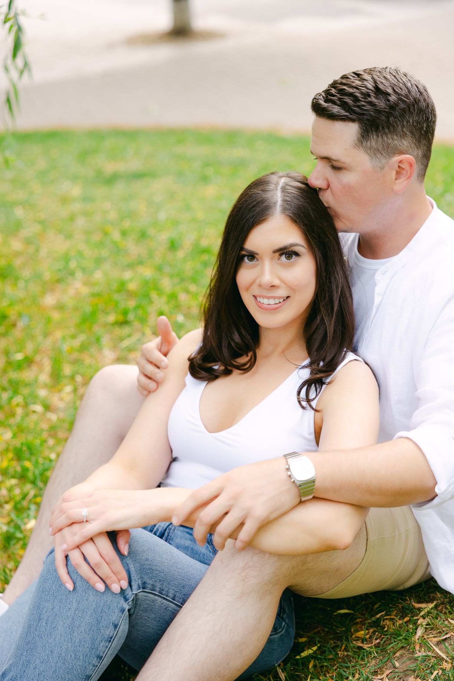 Romantic couple sitting together on grassy slope beneath willow branches.