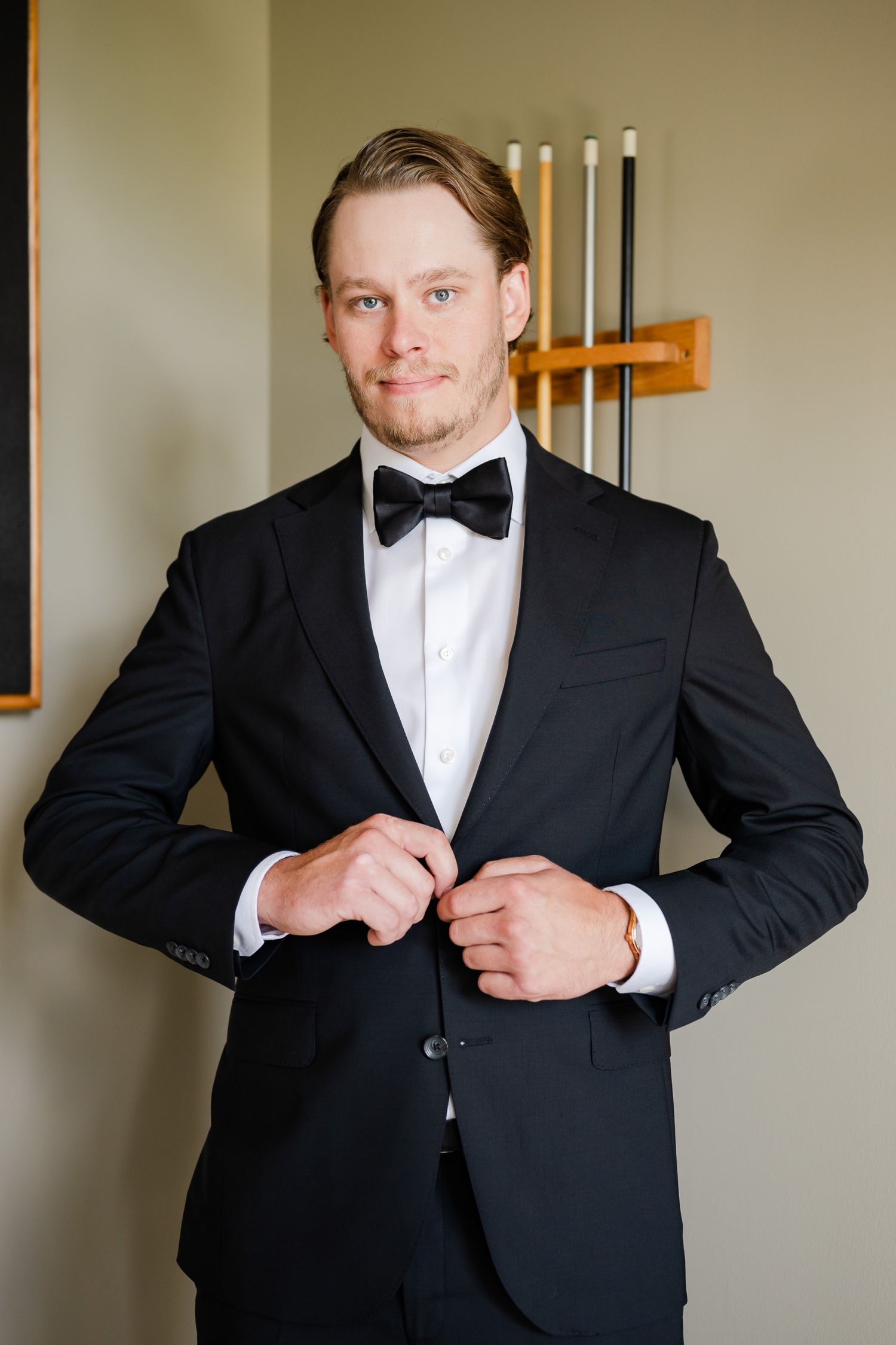 Person in black tuxedo and bow tie adjusting jacket lapels while standing near dartboard.