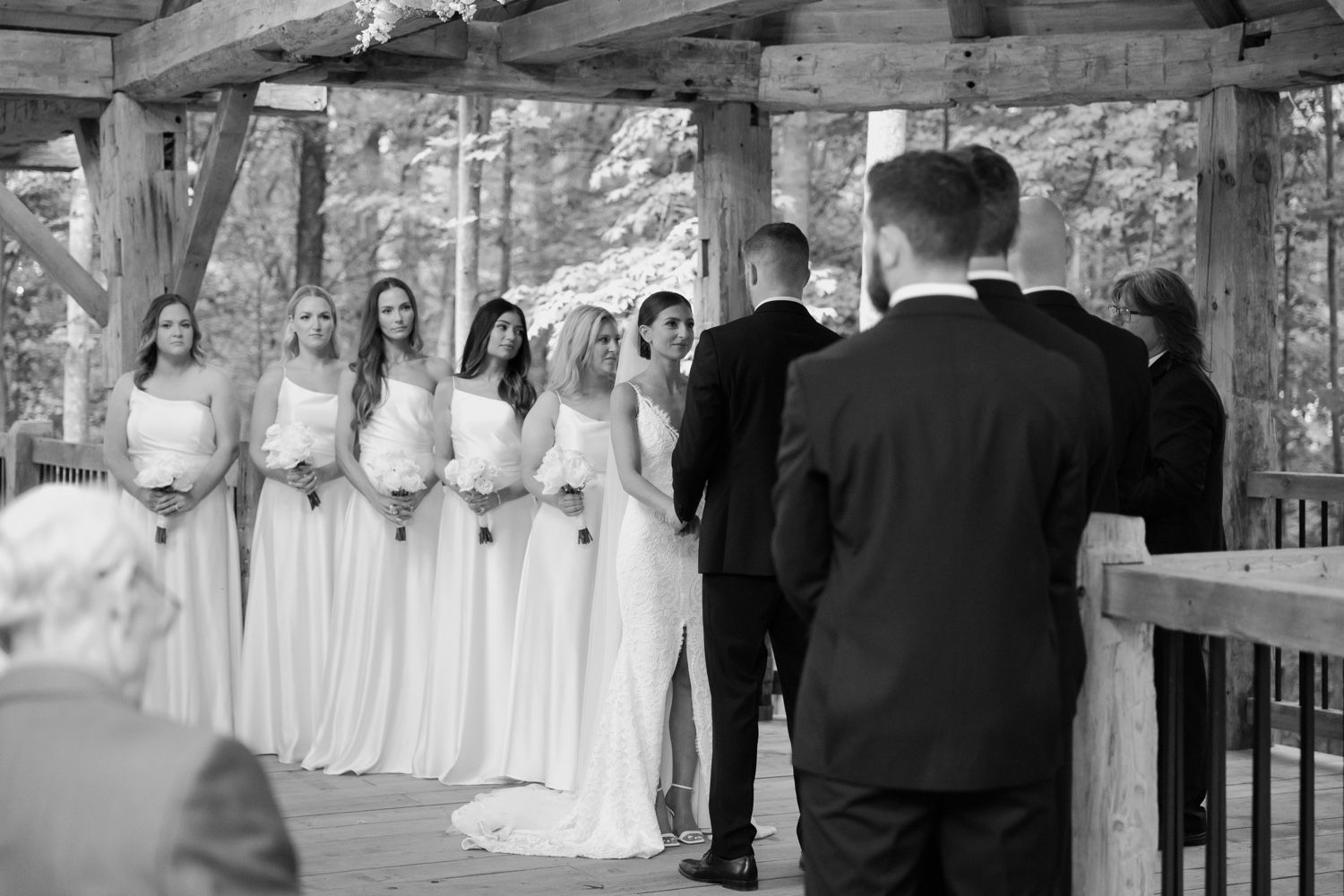 Black and white wedding ceremony photo with wedding party in rustic outdoor pavilion.