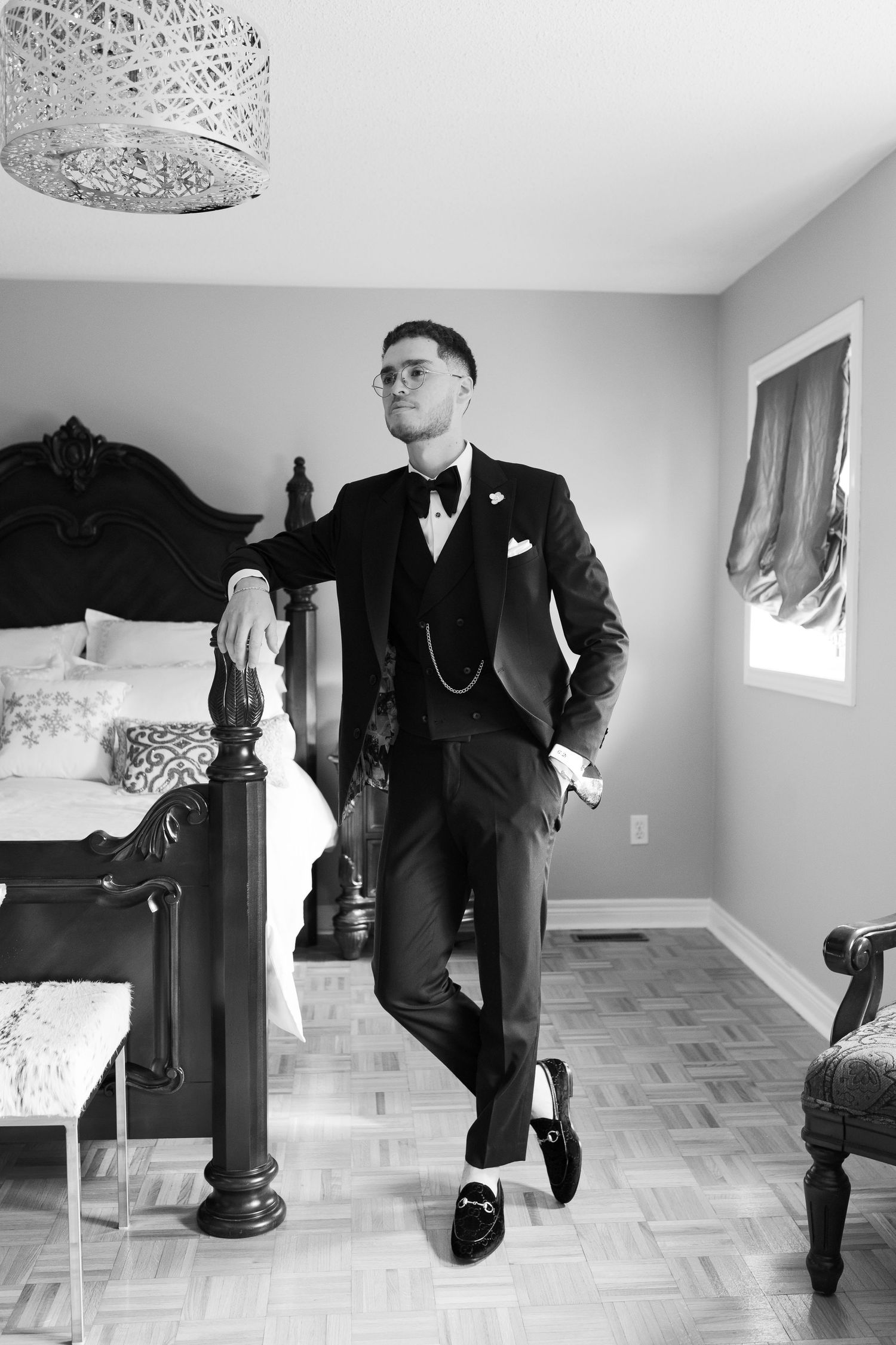 Black and white portrait of someone posed in a classic tuxedo against an ornate bedframe.
