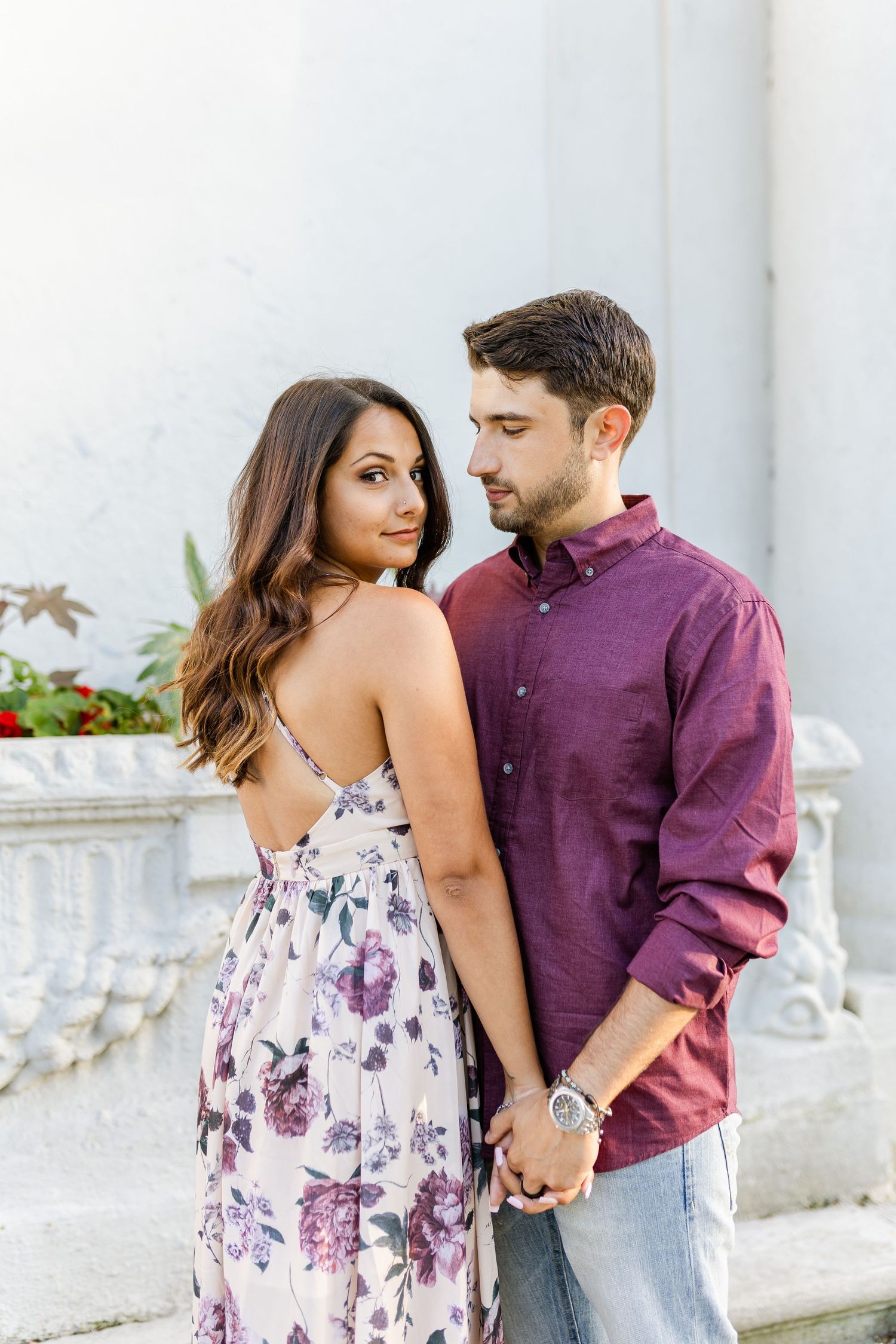A couple in burgundy and floral outfits poses intimately by a white architectural feature.