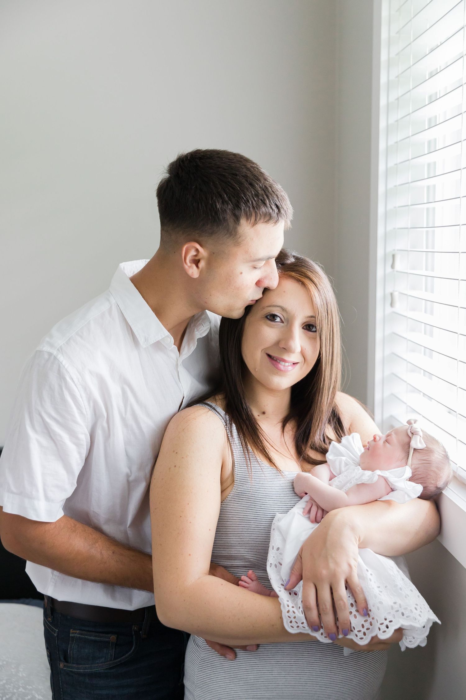 A tender family moment as parents cradle their newborn baby in white lace dress by a bright window.