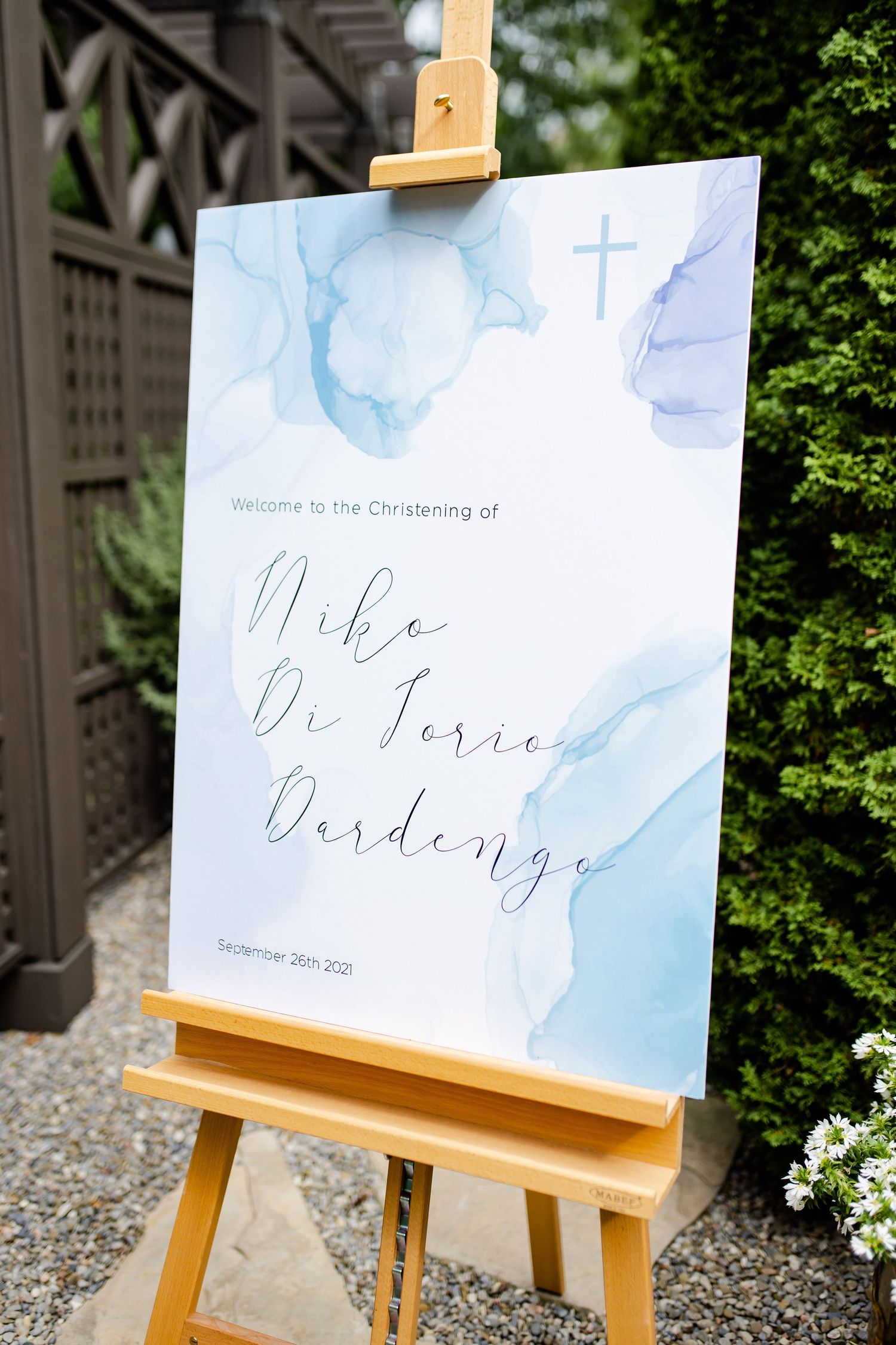 A blue watercolor christening invitation for Mike displayed on a wooden easel outdoors.