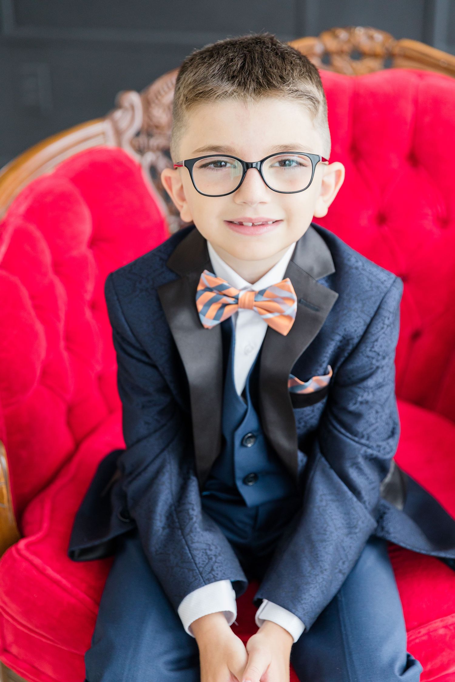 Young boy in navy suit and striped bow tie sits on red velvet chair.