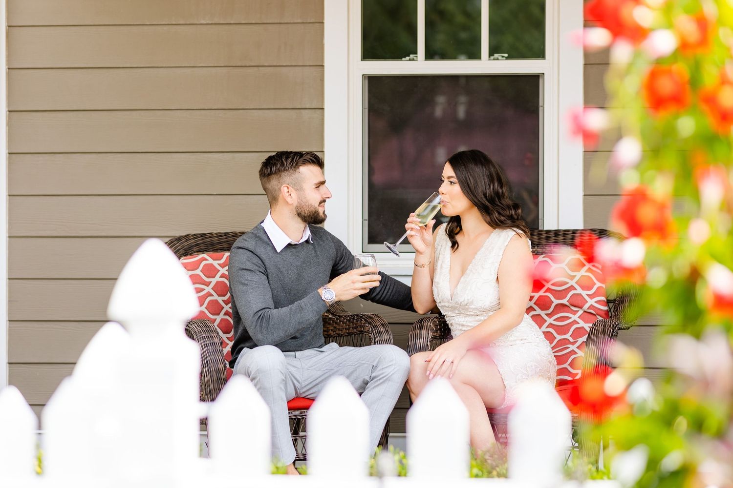 A couple shares drinks while sitting on a porch bench with red cushions.