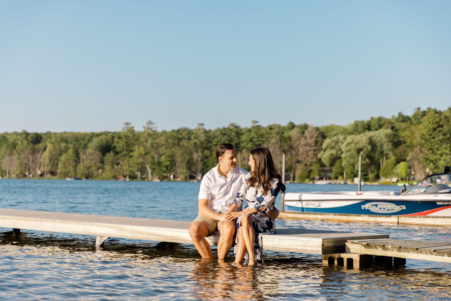 A couple sits together on a wooden dock overlooking a lake with boats in the background.