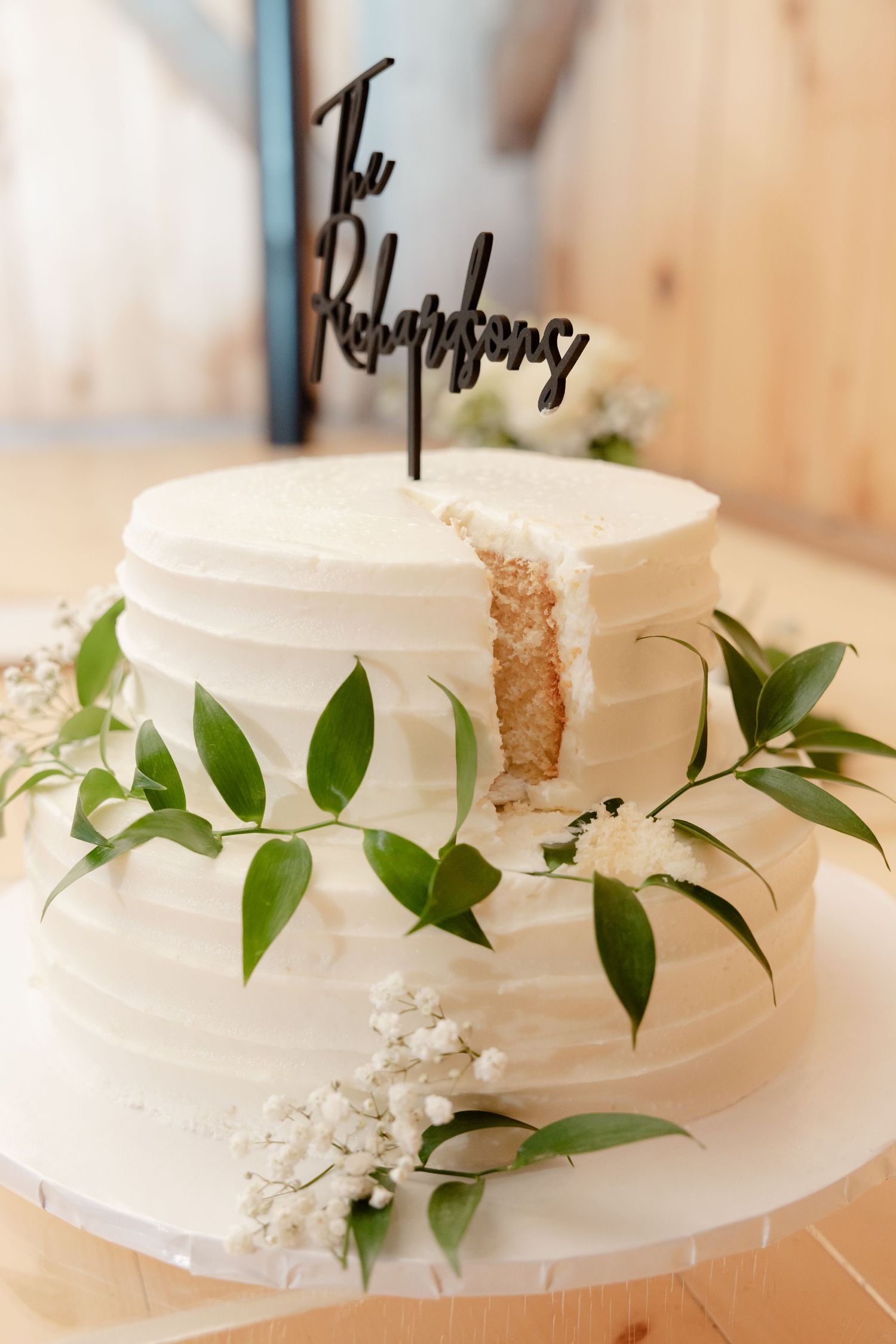 A white two-tier wedding cake decorated with fresh greenery and baby's breath on a wooden surface.