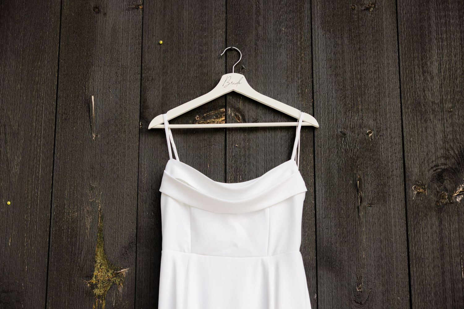 A white wedding dress hangs on a wooden hanger against dark wooden planks.