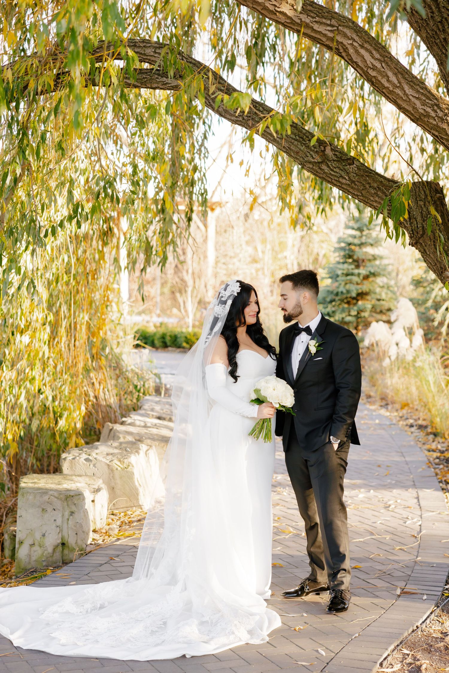 Newlyweds share an intimate moment under golden willow branches on a garden path.