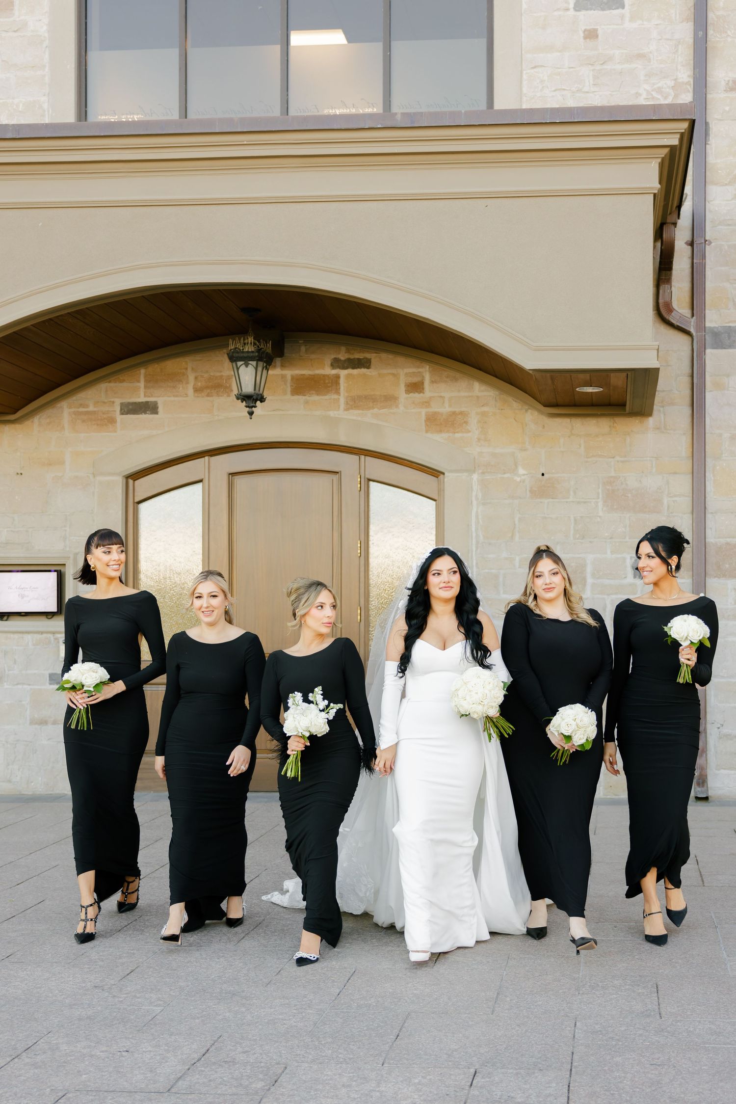 Wedding party poses in classic black bridesmaids dresses and white bridal gown against stone archway.
