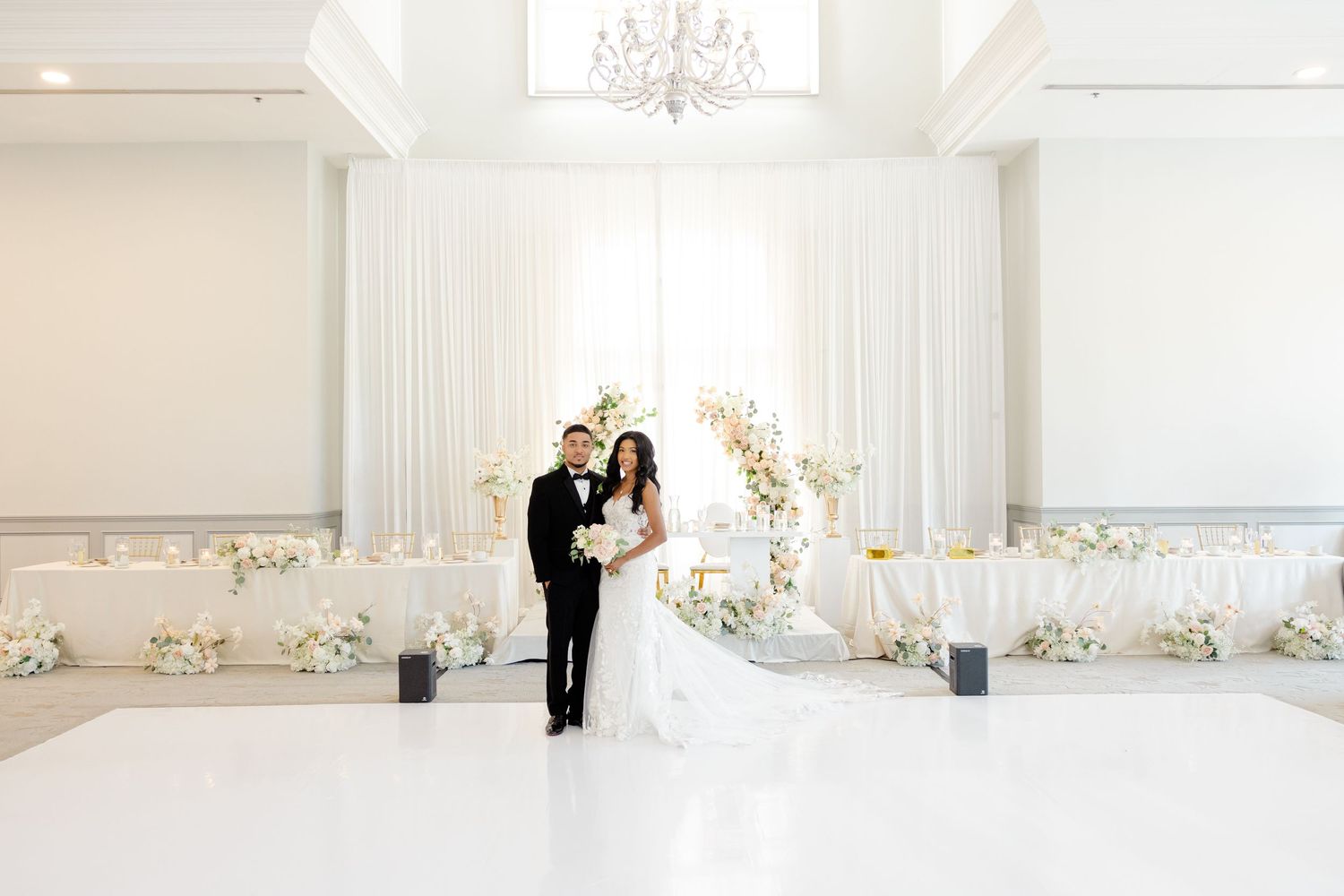 Wedding couple embracing in front of white drapes and floral arch in bright ballroom.
