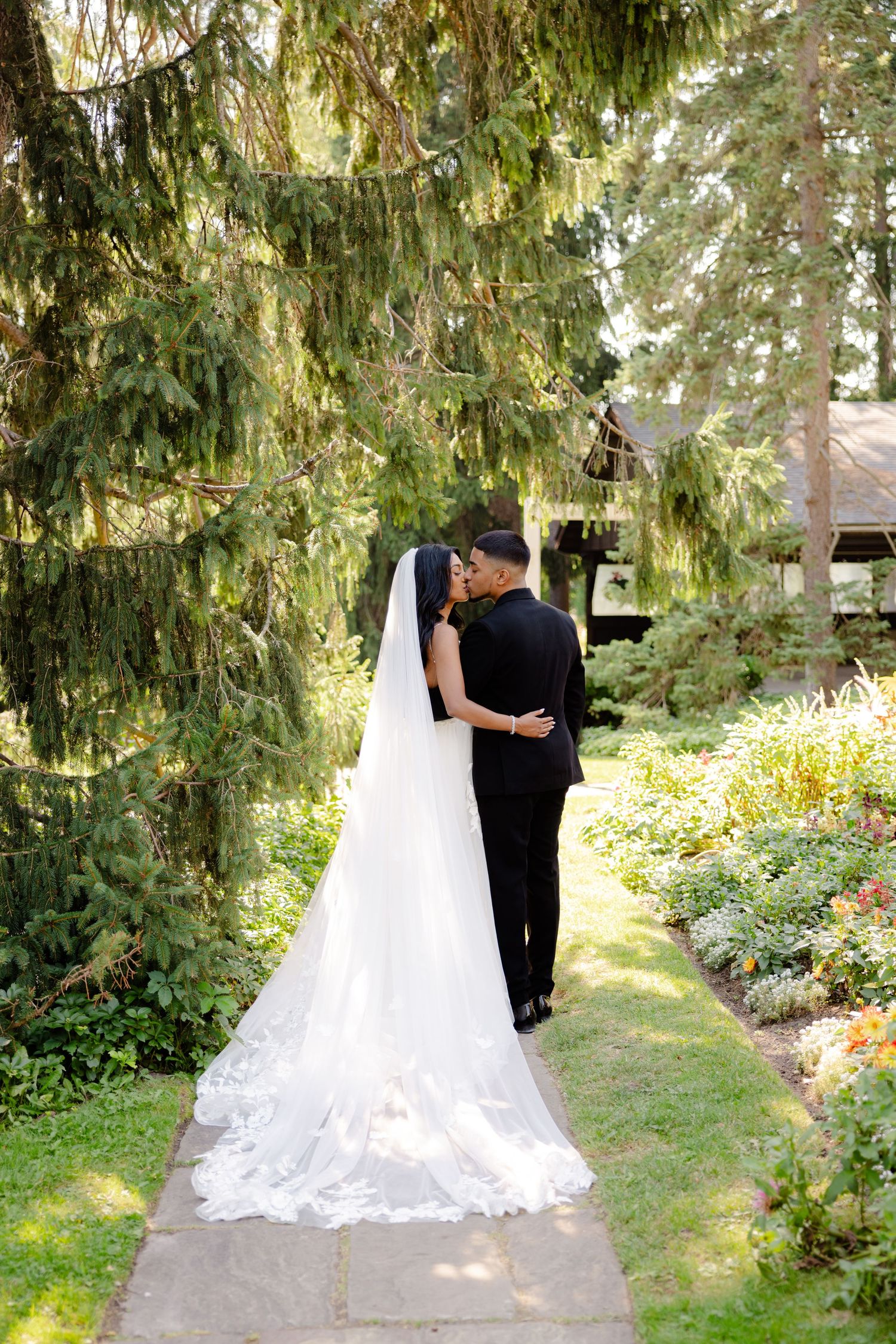Newlyweds share a tender moment on a garden path under sweeping evergreen branches.