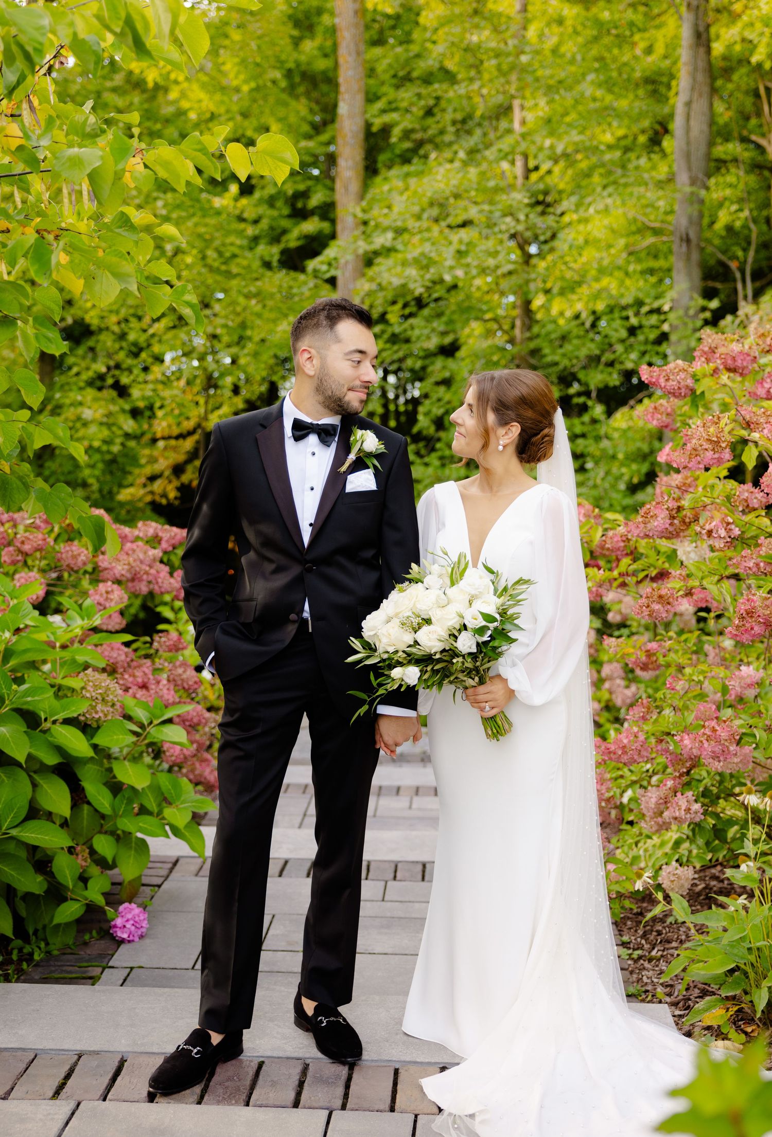 Joyful wedding couple standing on garden pathway surrounded by pink flowers and greenery.