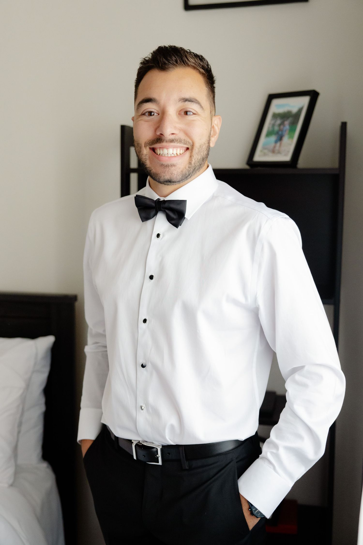 Groom wearing white dress shirt and black bow tie smiles brightly while getting ready in bedroom.