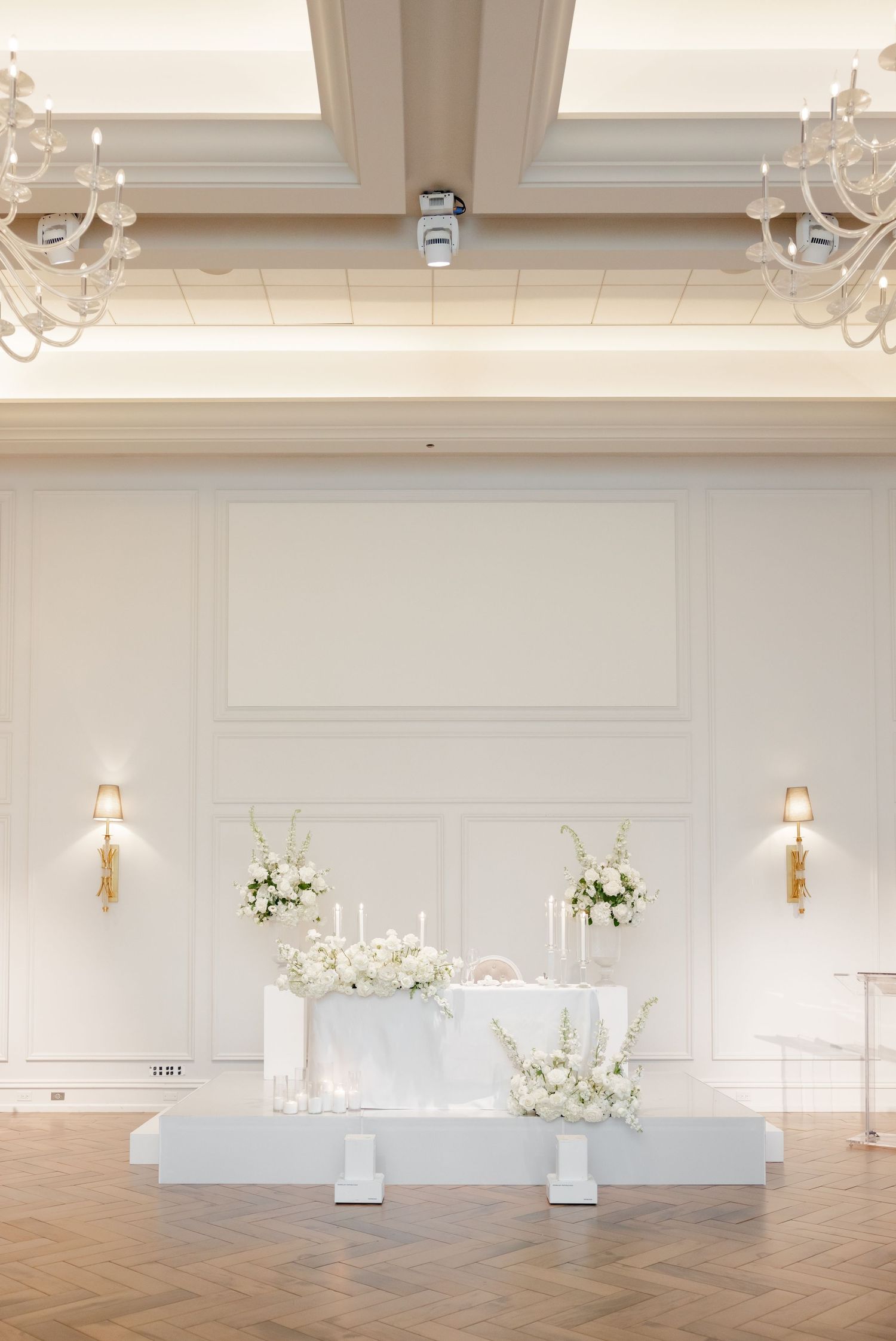 Elegant white wedding venue with crystal chandeliers and elevated floral head table display.