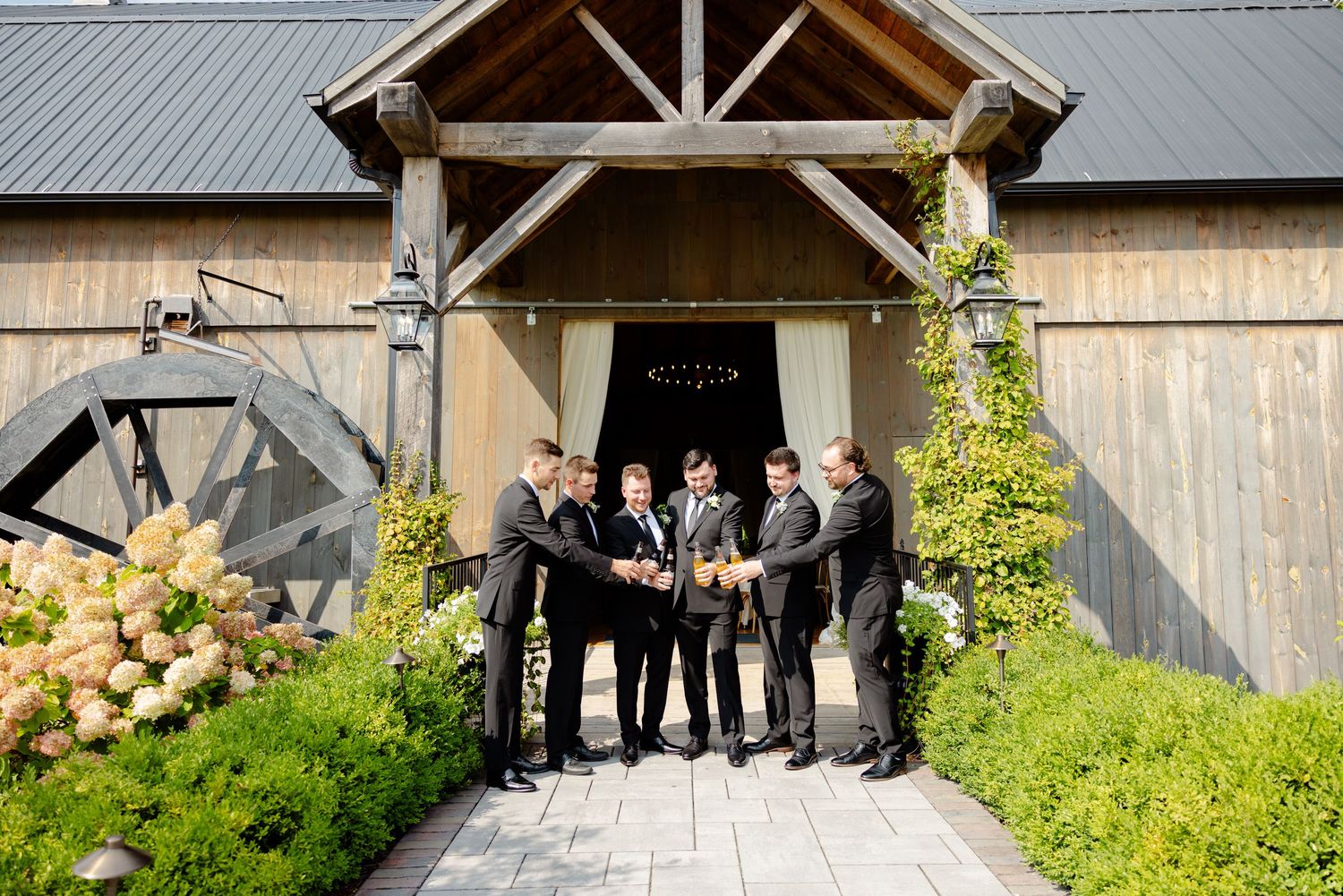 Groomsmen gather for a celebratory toast under a rustic barn entrance with climbing vines and hydrangeas.