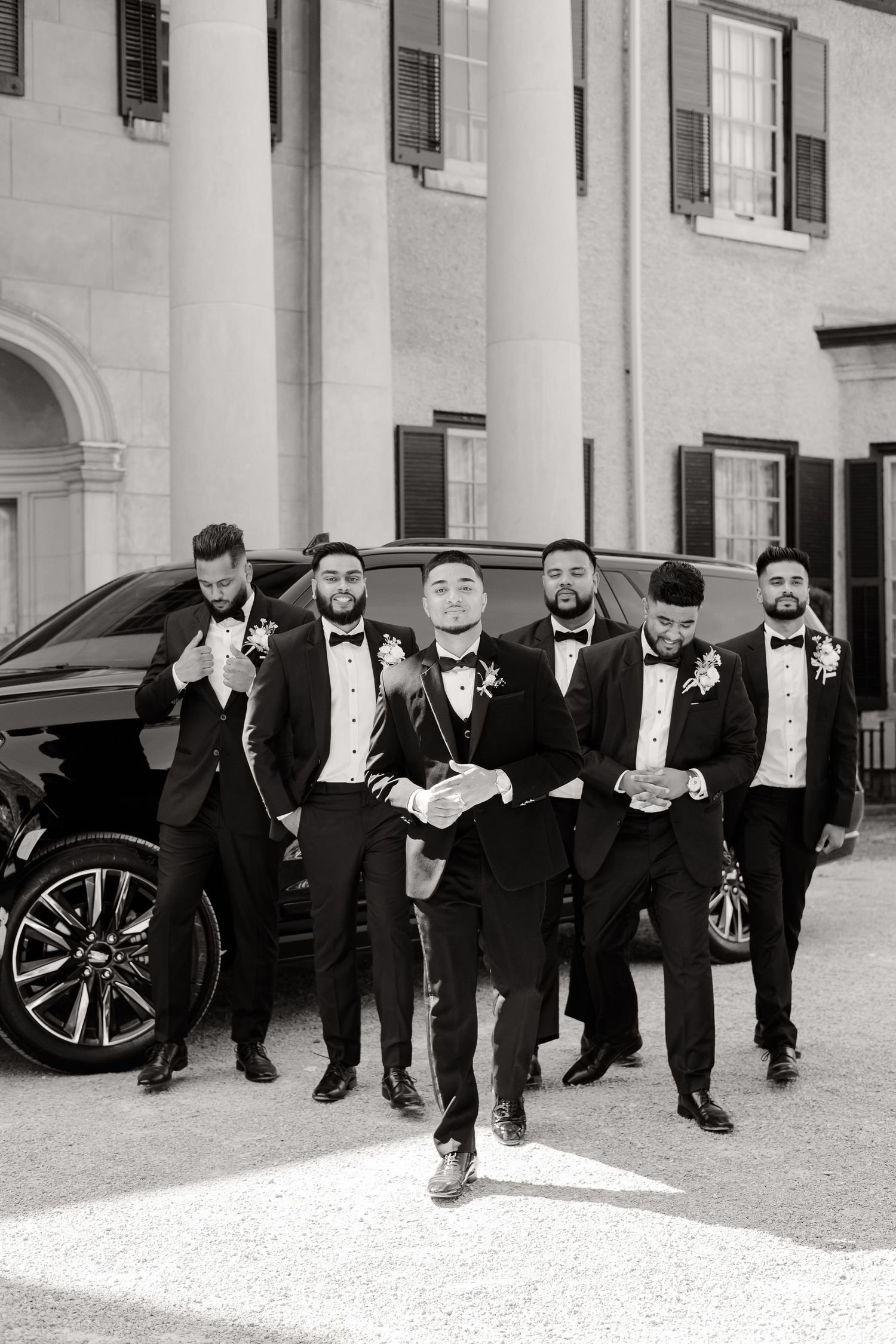 A groomsmen group in black tuxedos poses confidently in front of a black luxury vehicle and building with columns.