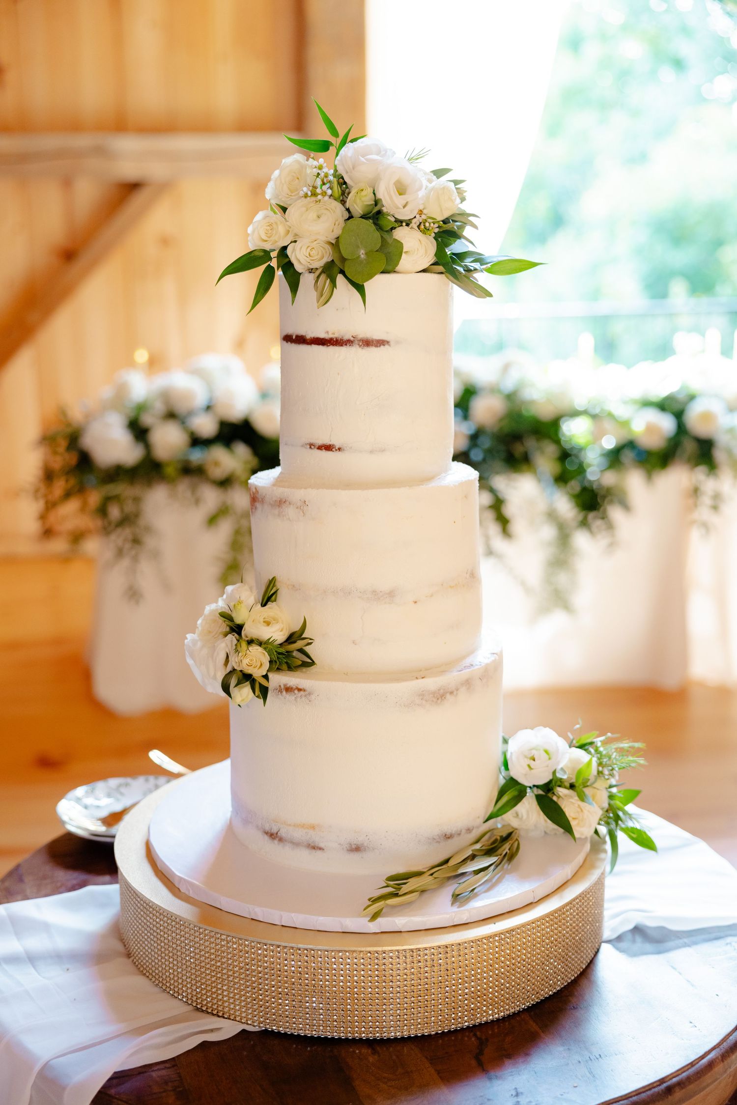 A four-tier semi-naked wedding cake decorated with white flowers and greenery on a gold base.