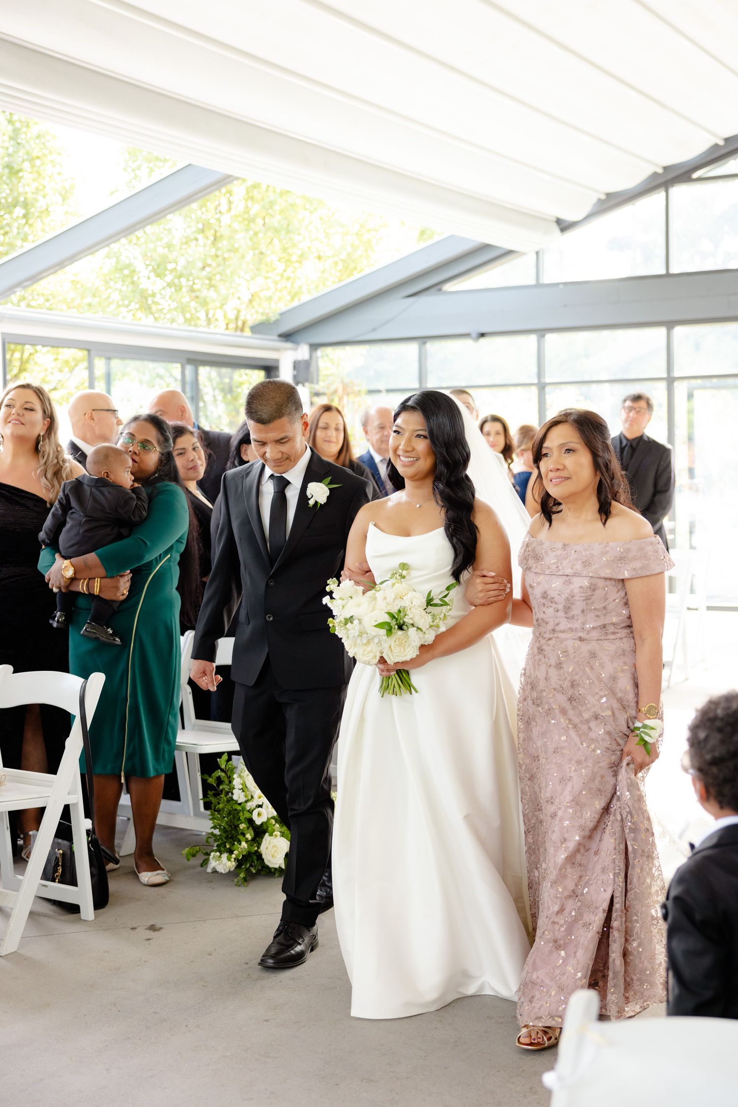 Wedding party walks between rows of white chairs in a sunlit venue with glass walls.