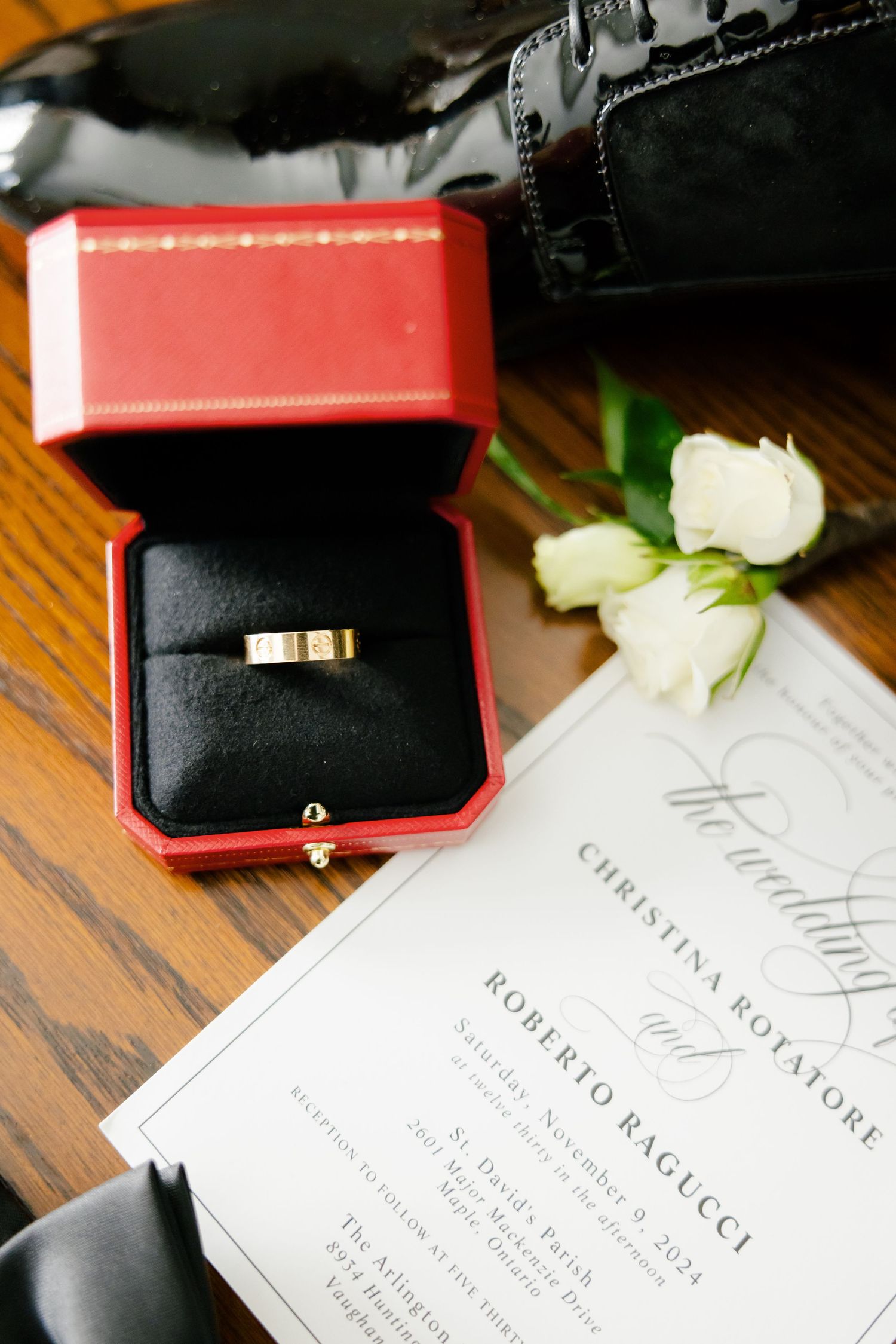 Wedding ring in red box beside white roses and elegant invitation on wooden table.