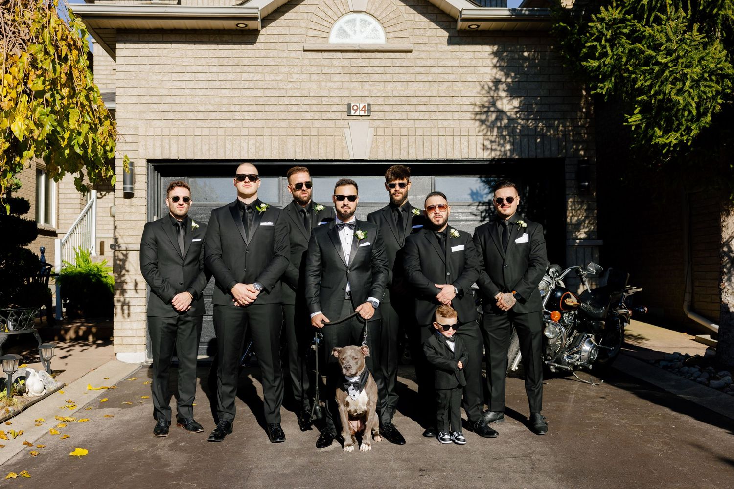 People in dark suits stand in formation with a dog on a driveway.