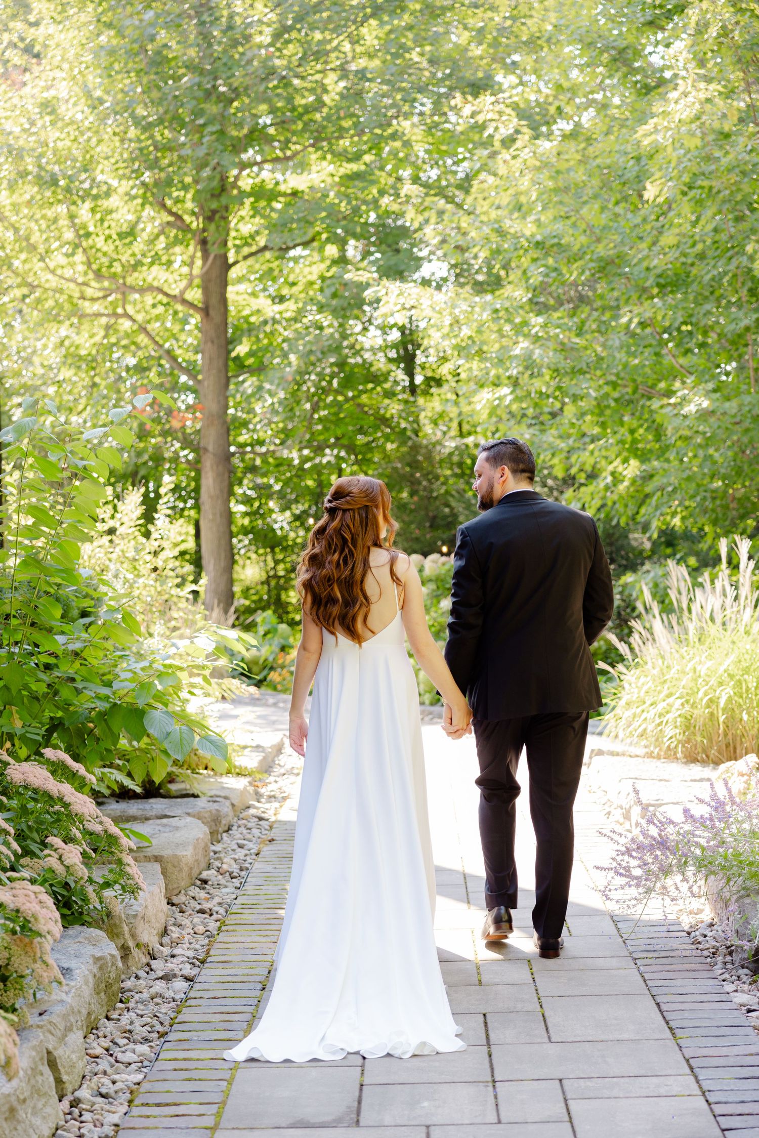 A couple walks hand in hand down a stone garden path surrounded by lush greenery and flowers.