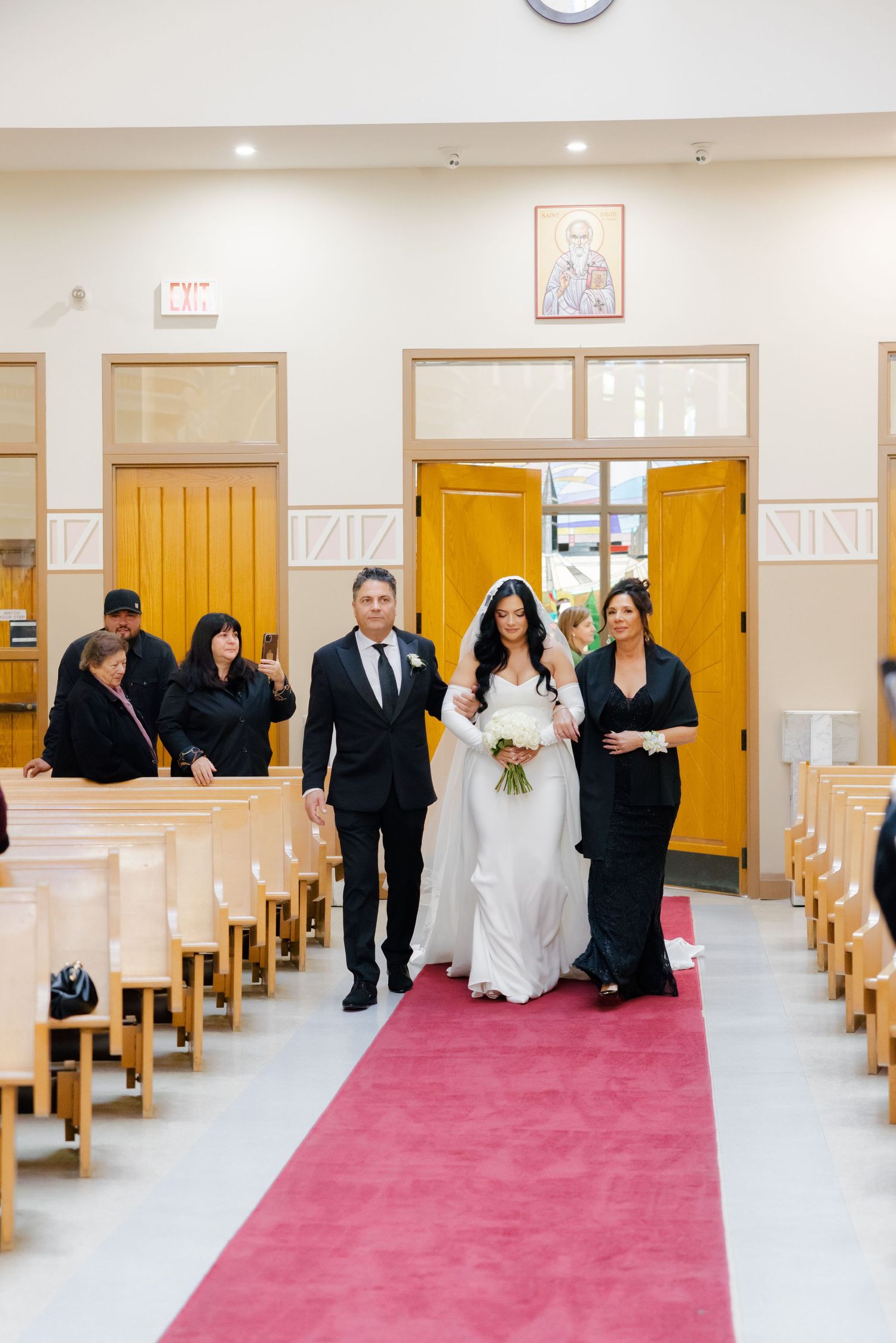 A wedding procession walks down a church aisle with yellow doors and wooden pews.
