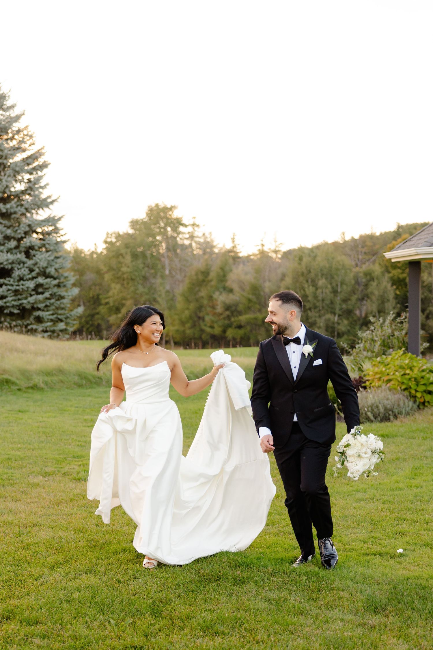 Couple walking on grass with flowing wedding dress and evergreen trees in background.