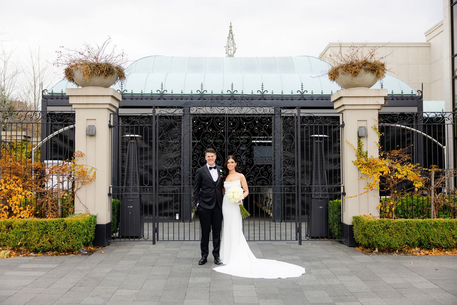 Bride and groom pose in front of an elaborate iron gate entrance with decorative columns.