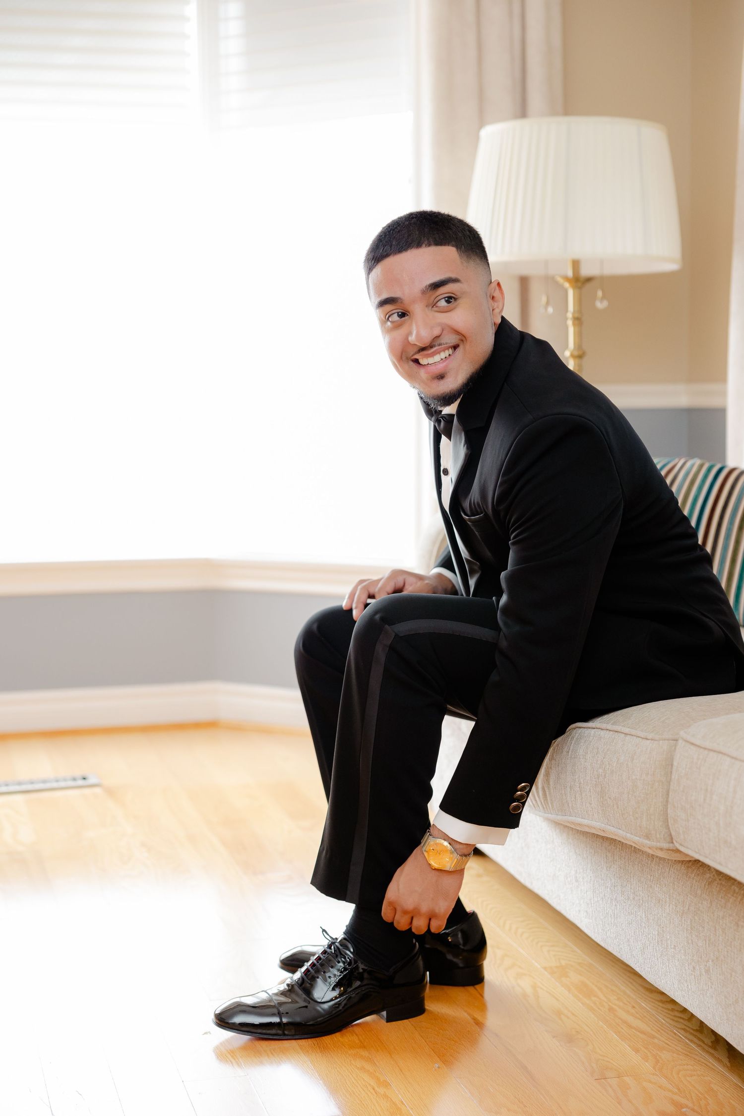 Groom in black tuxedo sits on white couch while getting ready for wedding.