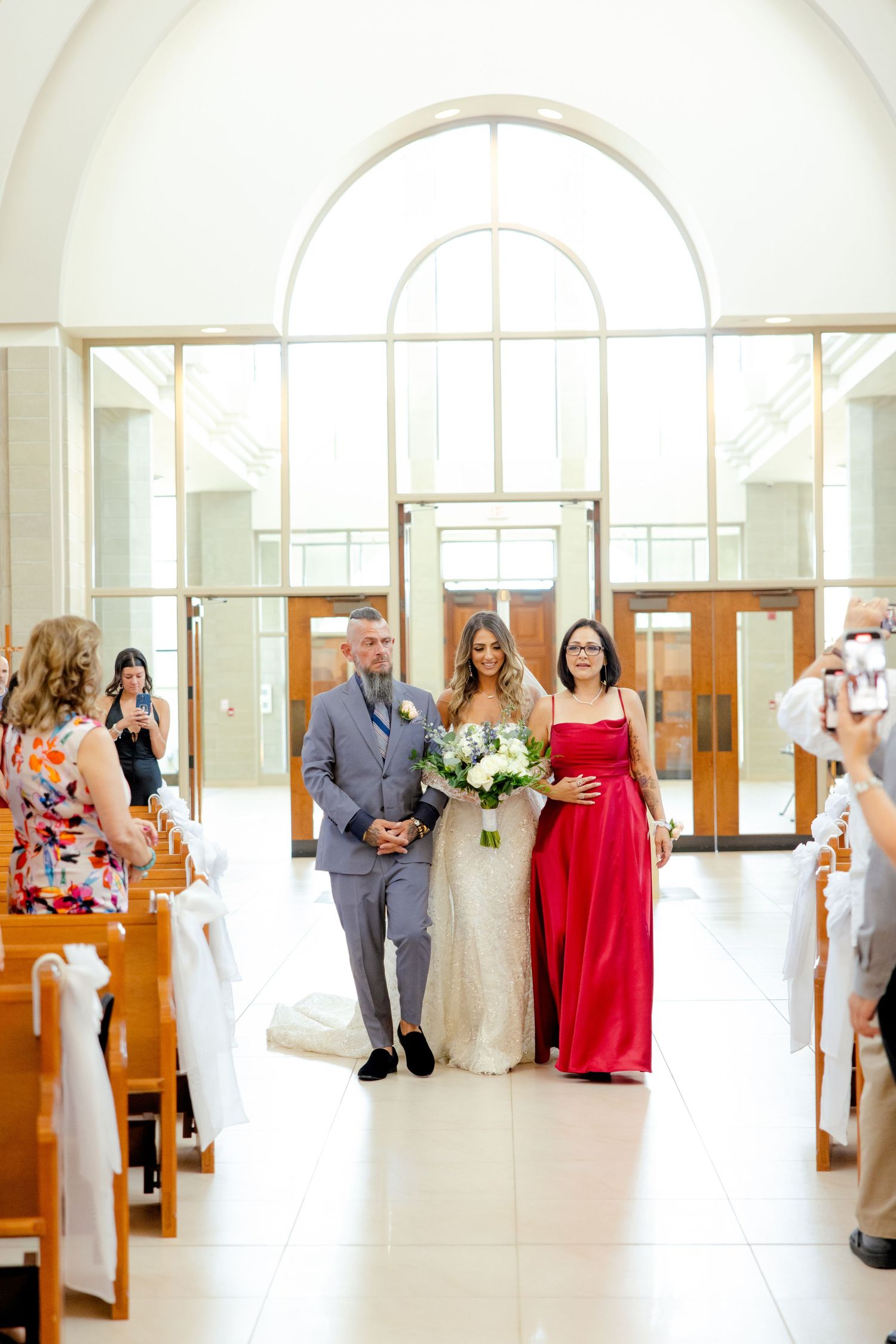 A wedding party walks down the aisle in a bright modern church with high arched windows.