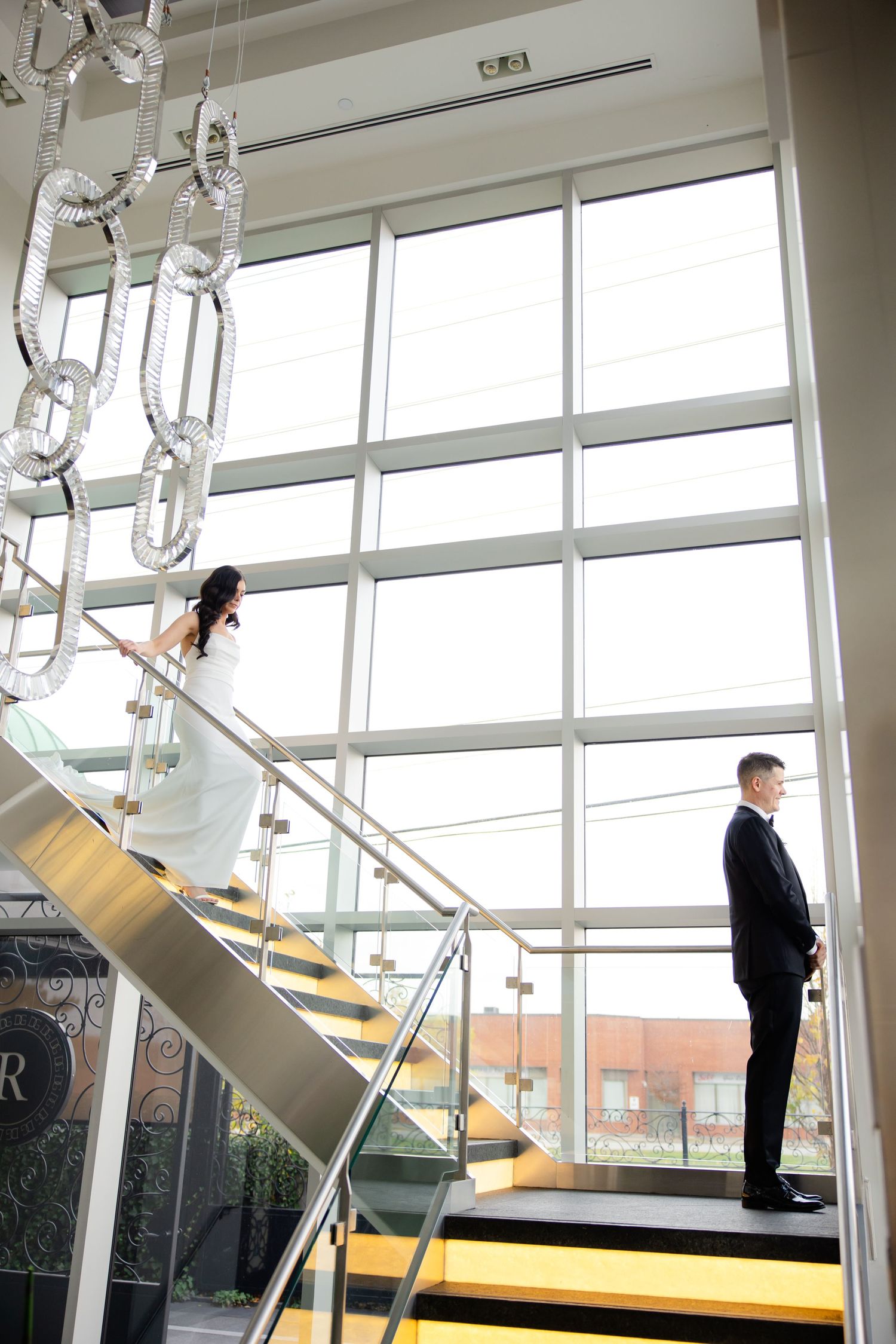 Wedding couple poses separately on yellow-accented stairs with dramatic floor-to-ceiling windows and crystal lighting.