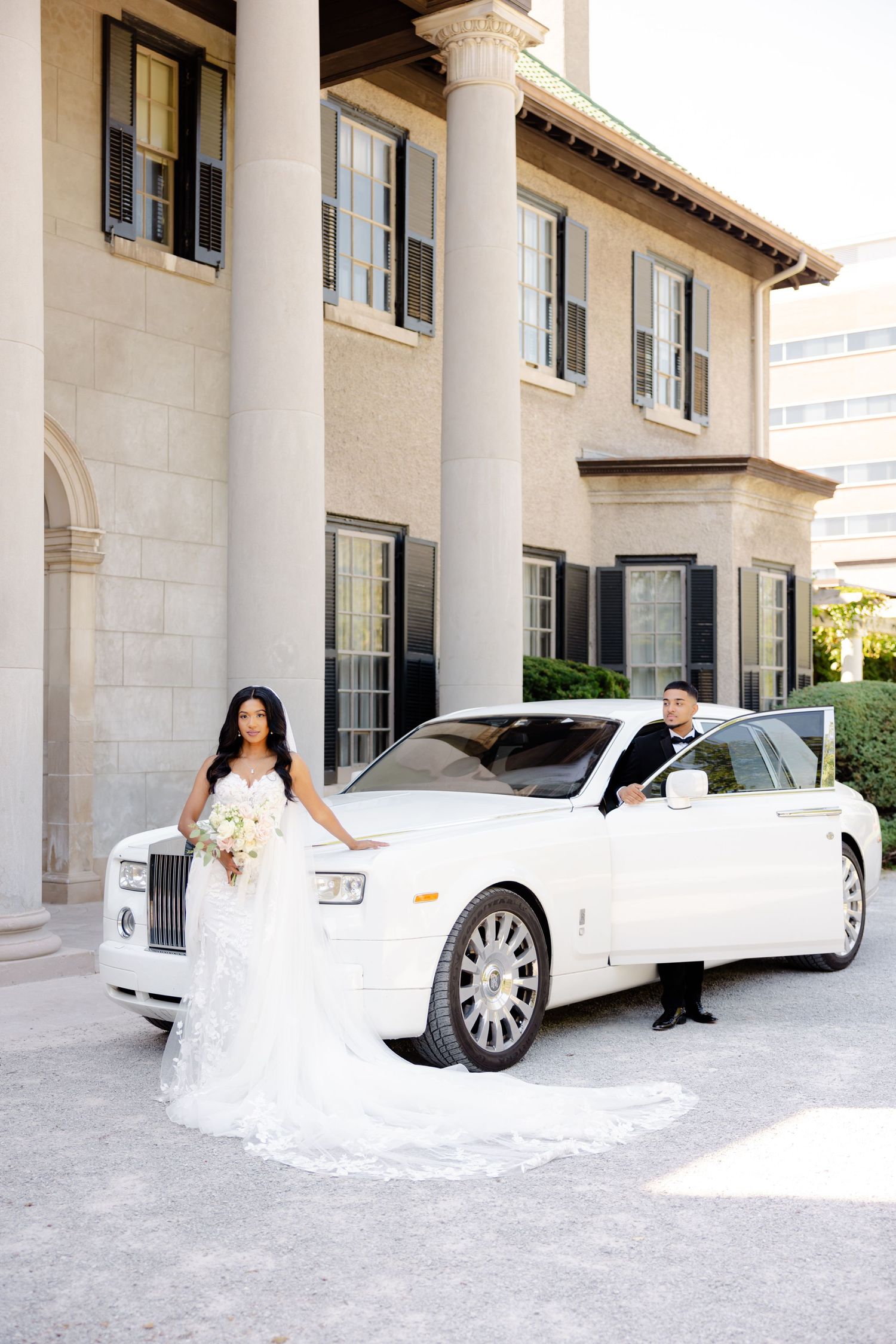 Newlyweds pose with a luxurious white Rolls-Royce in front of an elegant building with columns.