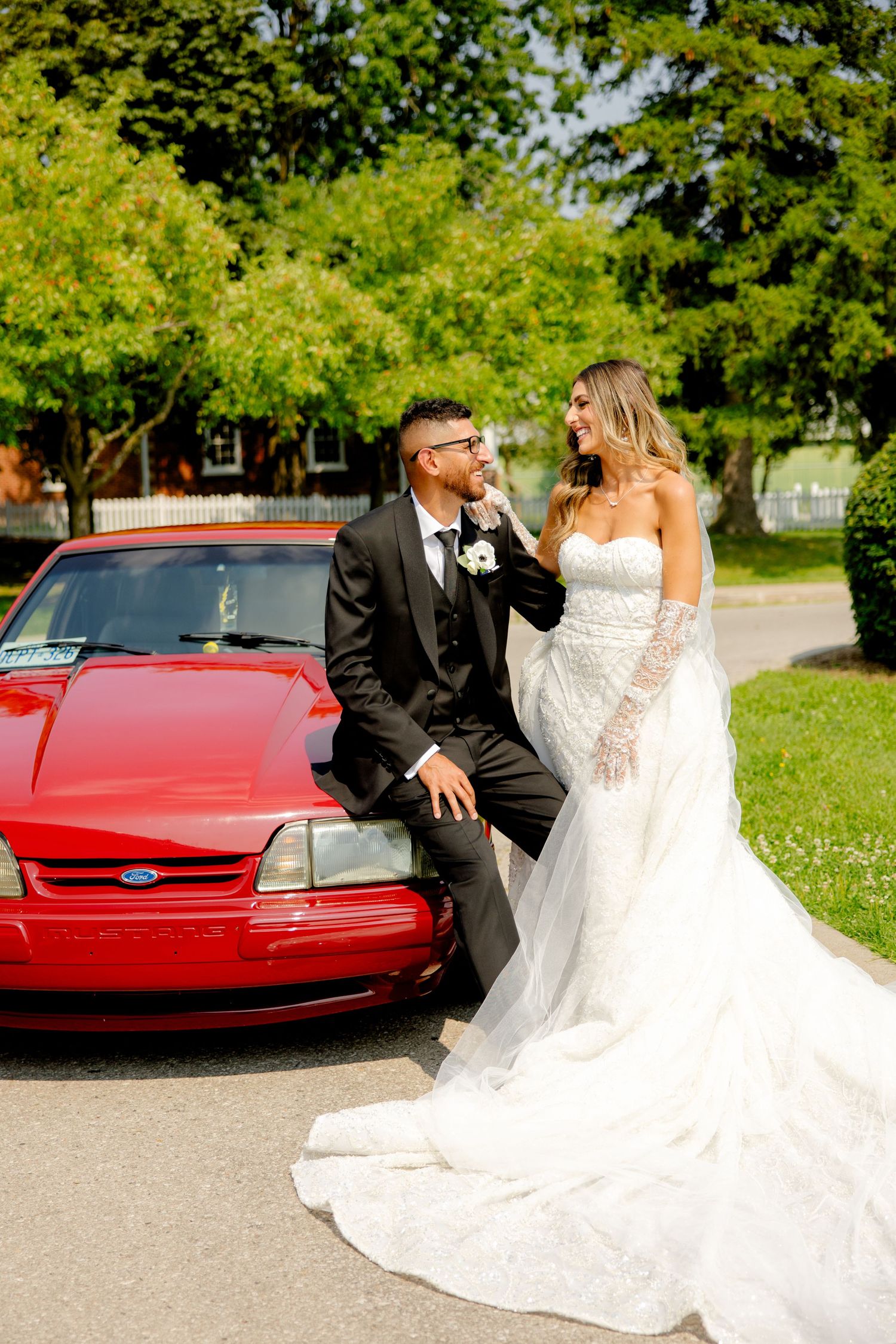 Romantic wedding photo with couple posed against their red car in a park setting.