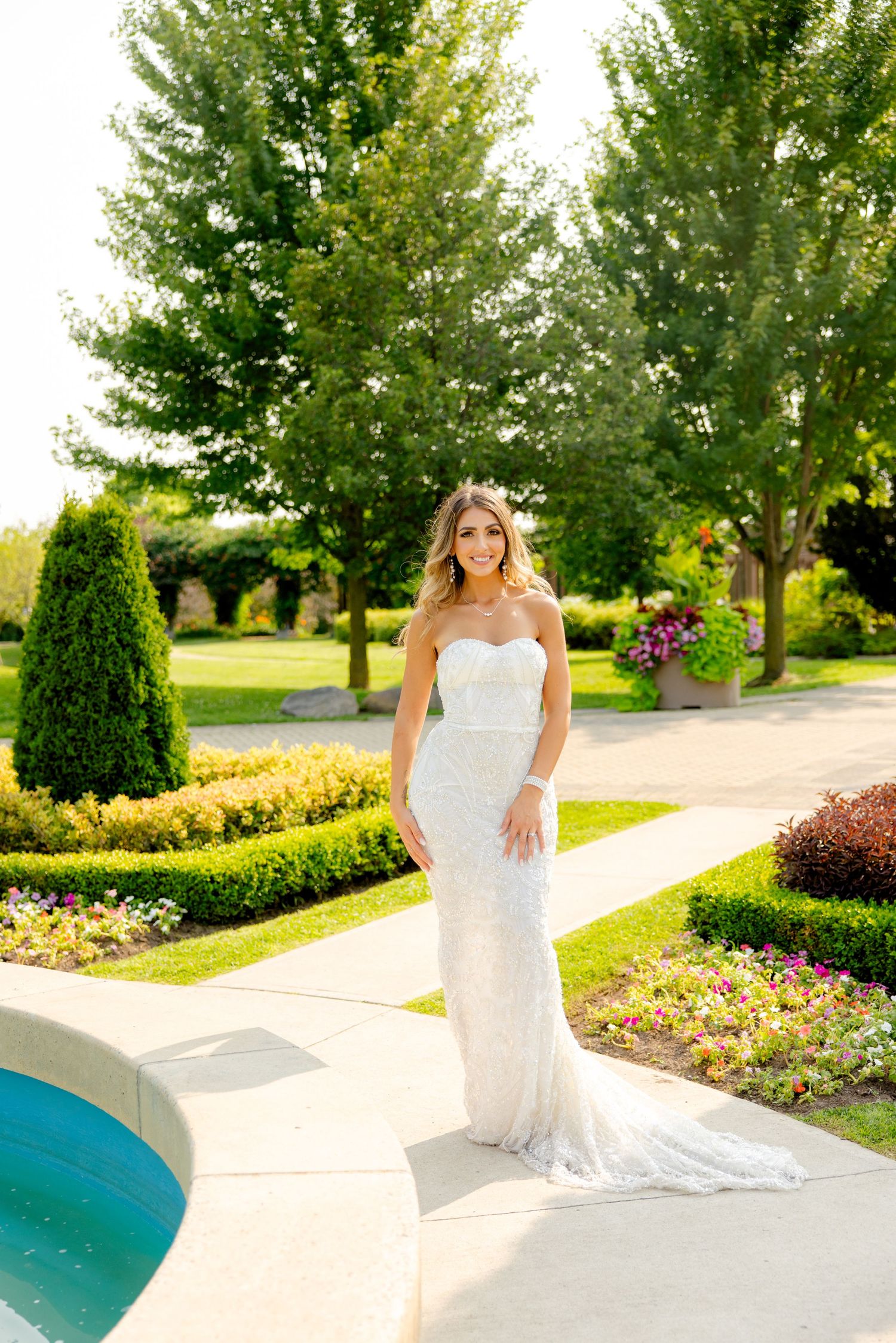 Bride in fitted white dress stands by pool in beautifully landscaped garden setting.