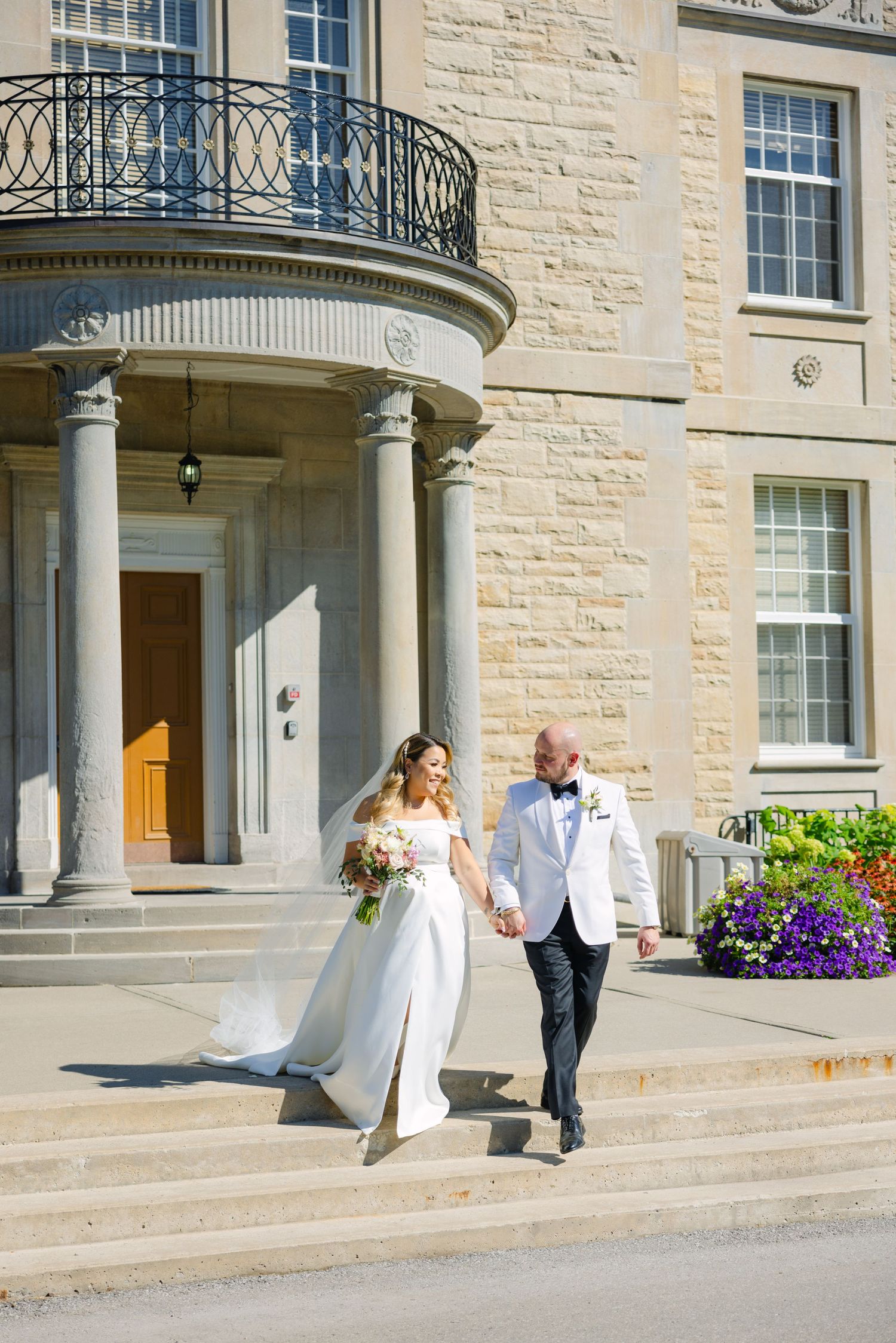 A wedding couple descends stone stairs of an elegant limestone building with columns and wrought iron balconies.