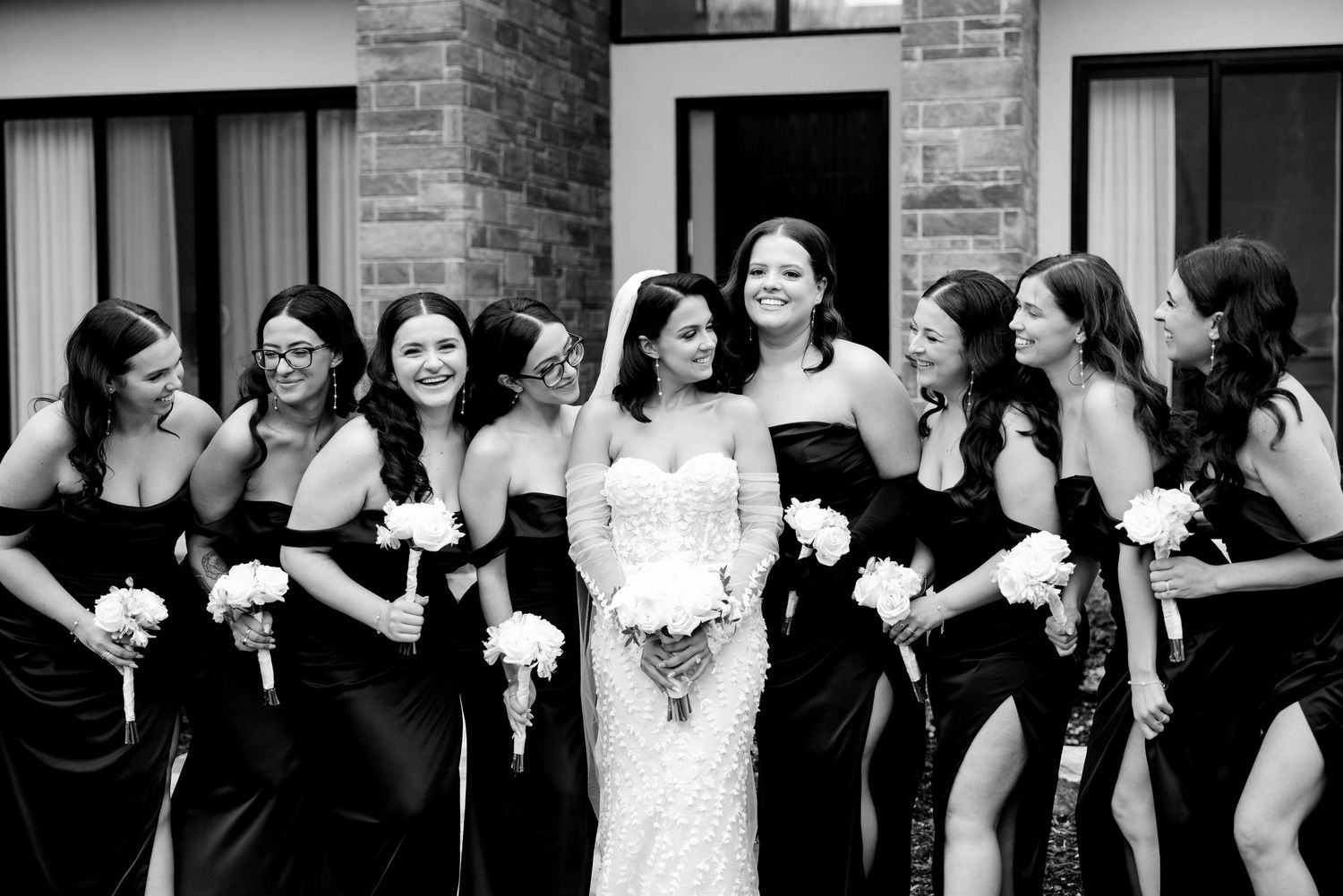 Bridesmaids in black dresses pose together on stone steps with the bride in white holding bouquets.
