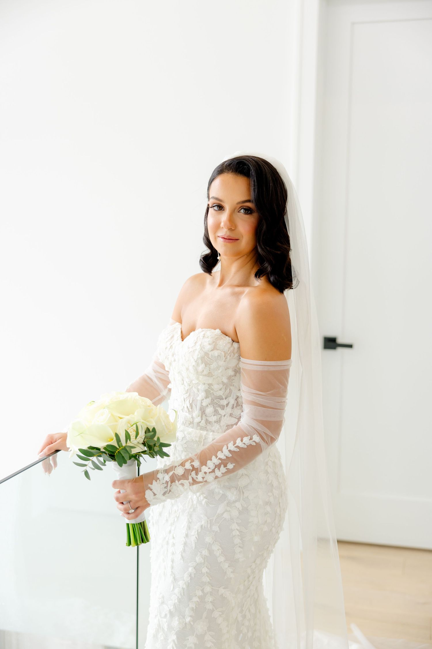 Bride poses elegantly in a fitted lace wedding gown with sheer sleeves while holding a classic white bouquet.