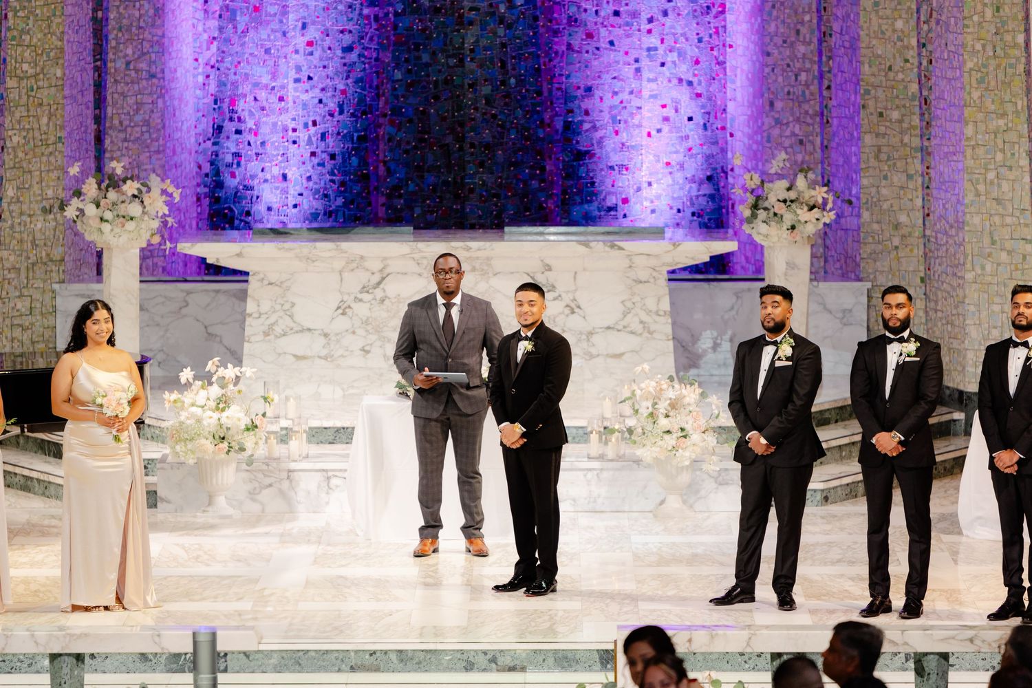 Wedding ceremony in progress at elegant church altar with purple lighting and white floral arrangements.