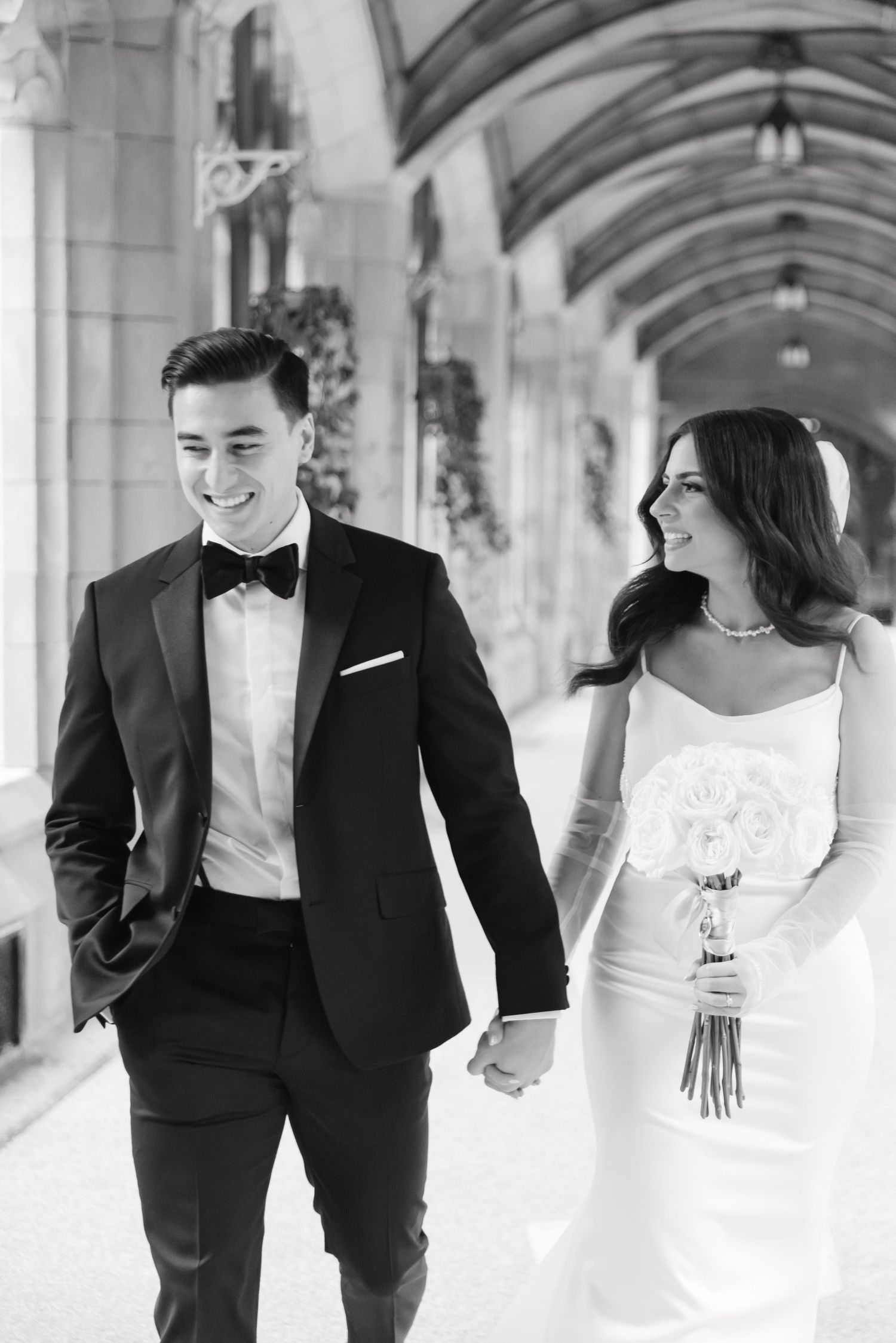 Black and white photo of a wedding couple walking and smiling together through an archway.