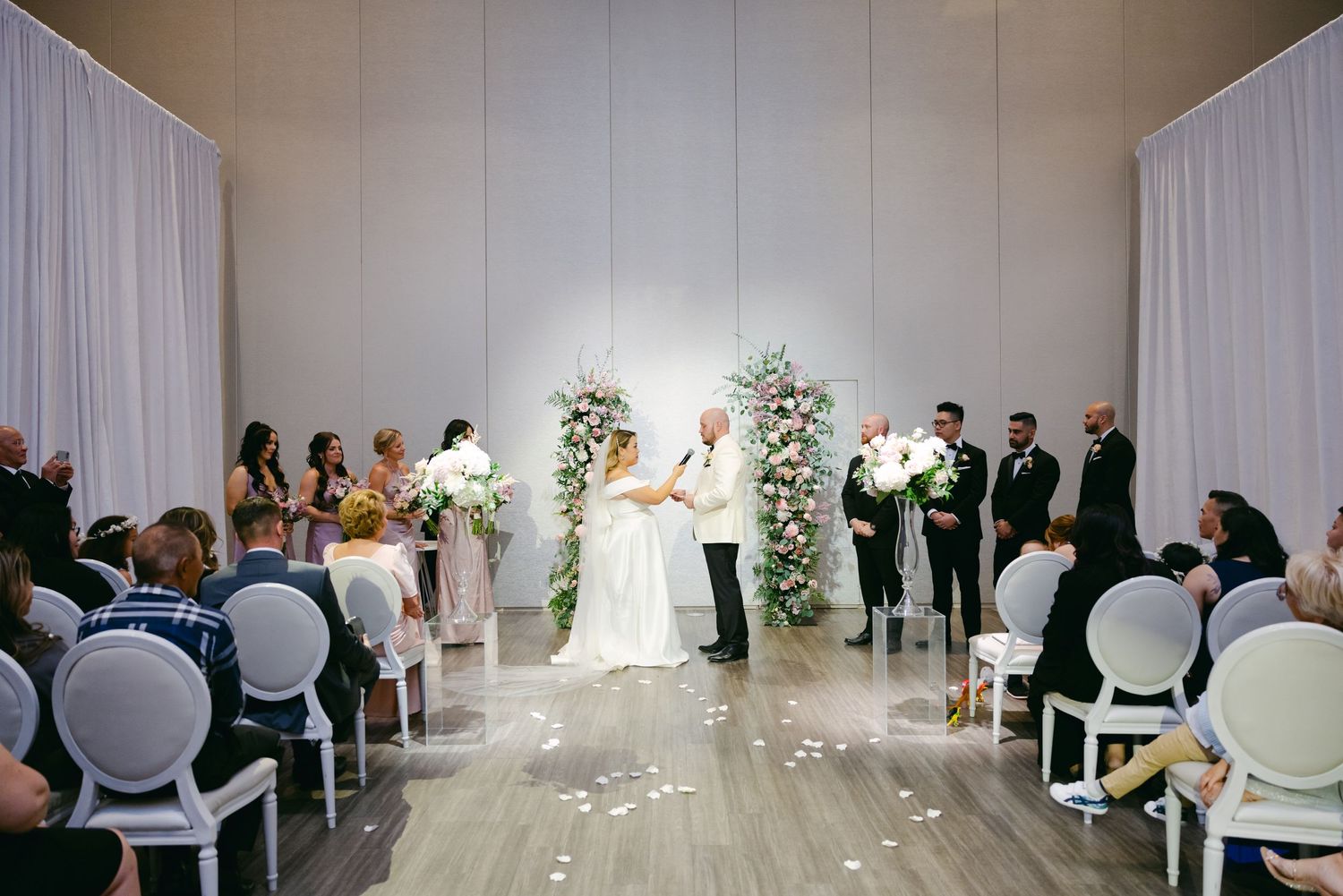 Wedding ceremony in elegant white room with floral arch and white chairs for guests.