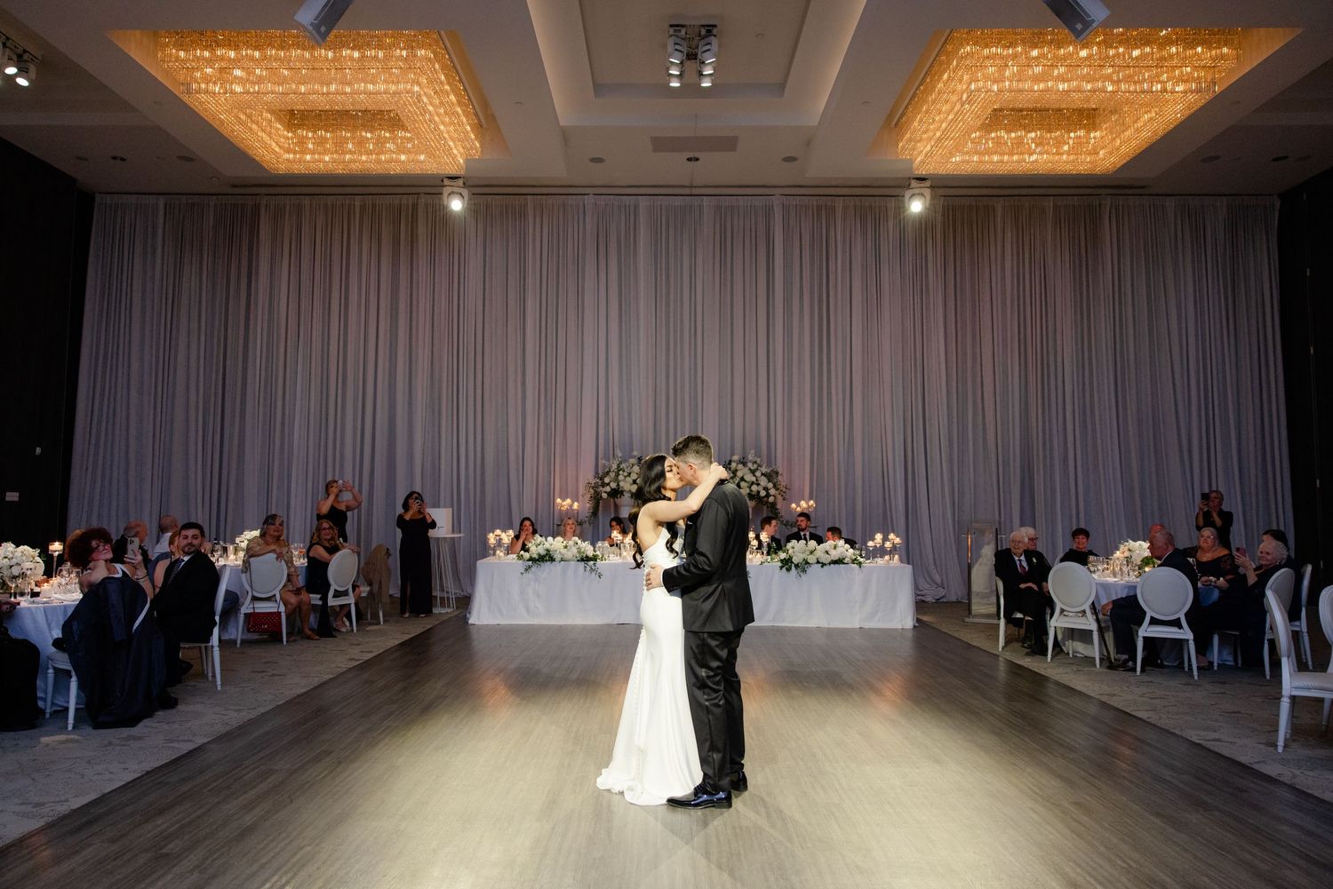 Newlyweds share an intimate dance moment in an elegantly lit ballroom with guests looking on.