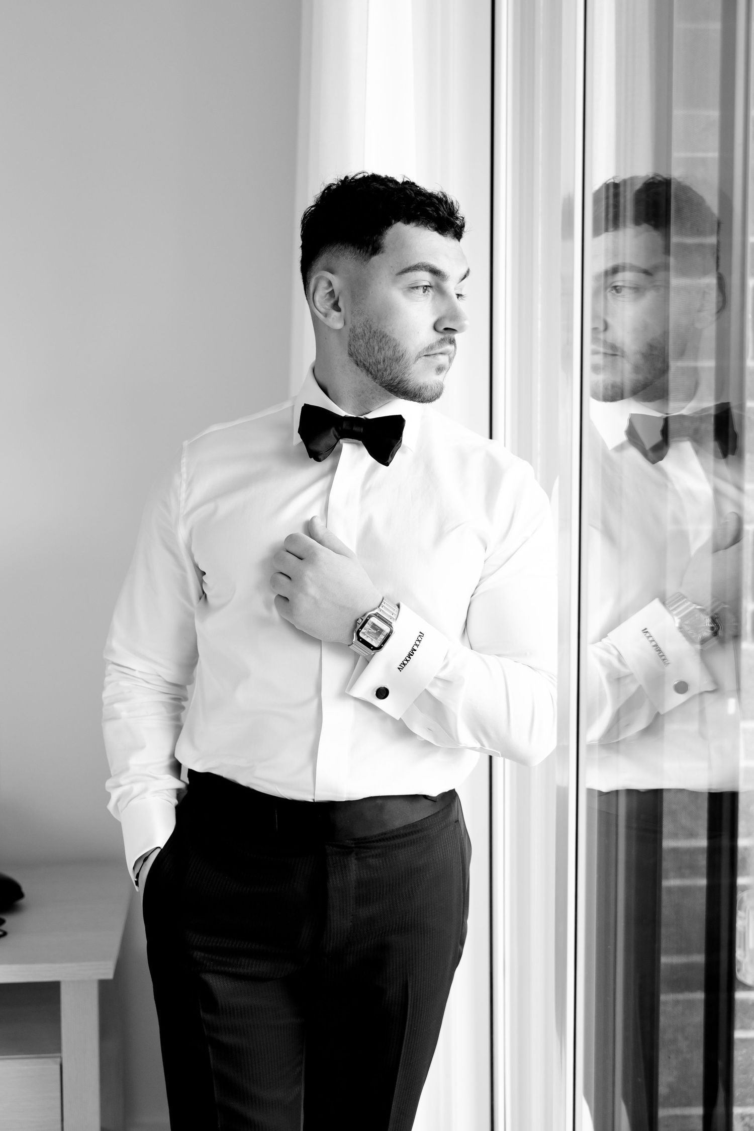 A formal attire featuring a crisp white shirt, black bow tie and sleek dress pants near a window.