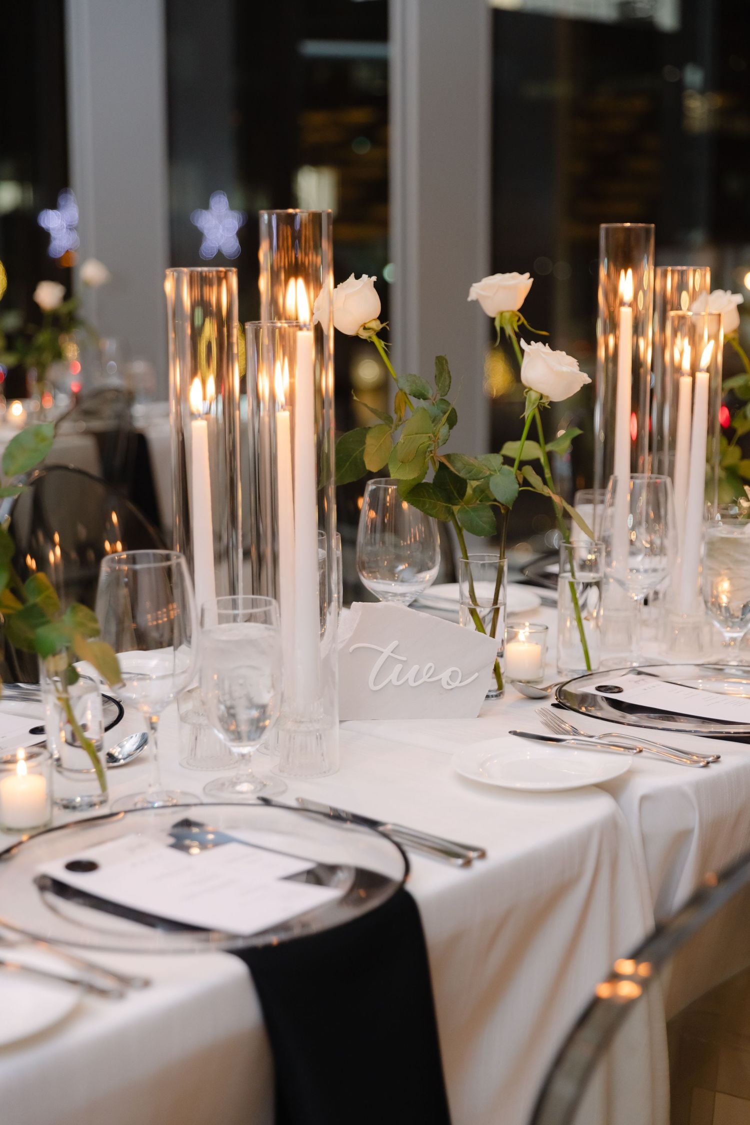 Wedding reception table with glass candle holders, white roses, and modern place settings.