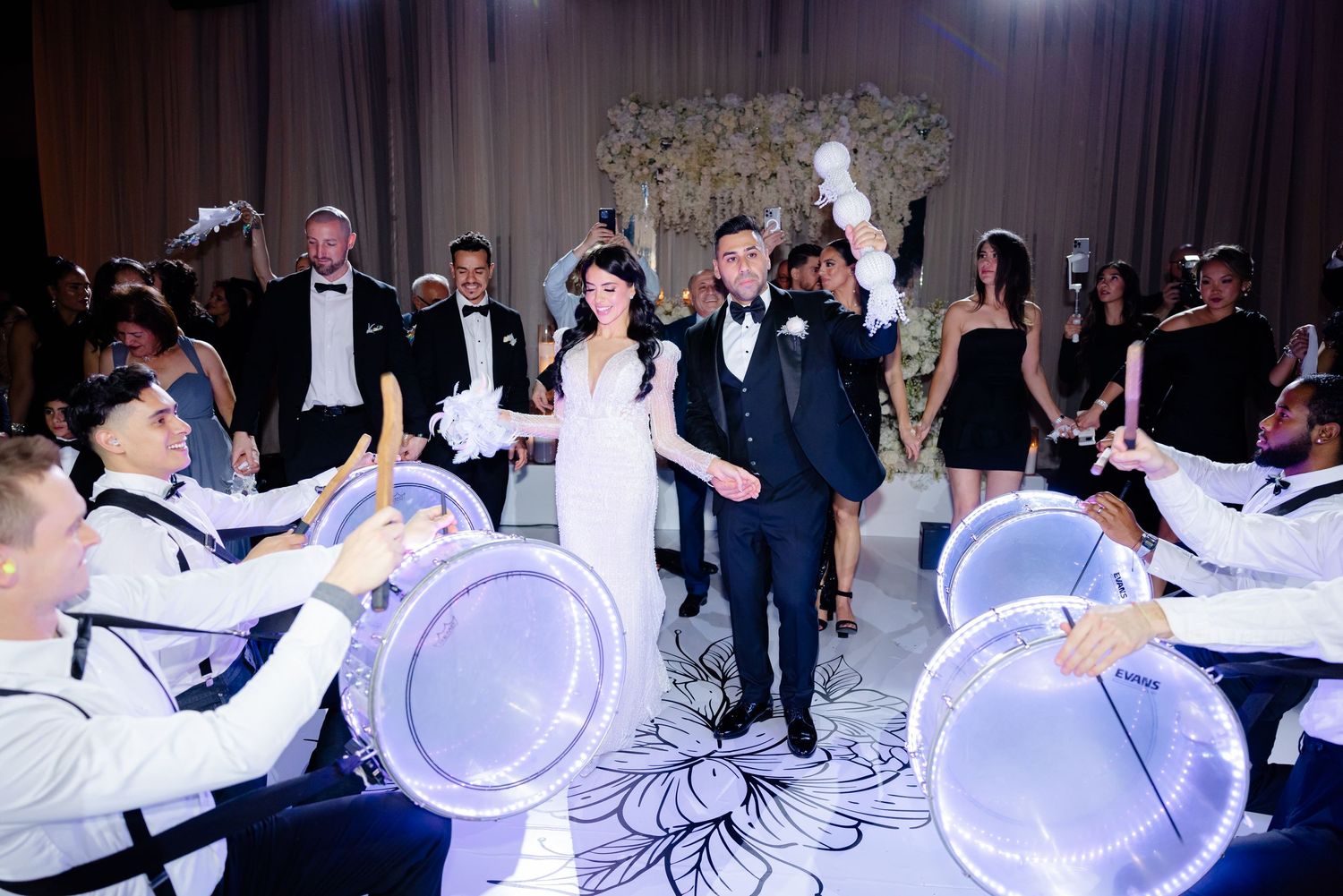 Elegant wedding celebration with LED-lit drummers performing on a patterned dance floor with white floral decor.