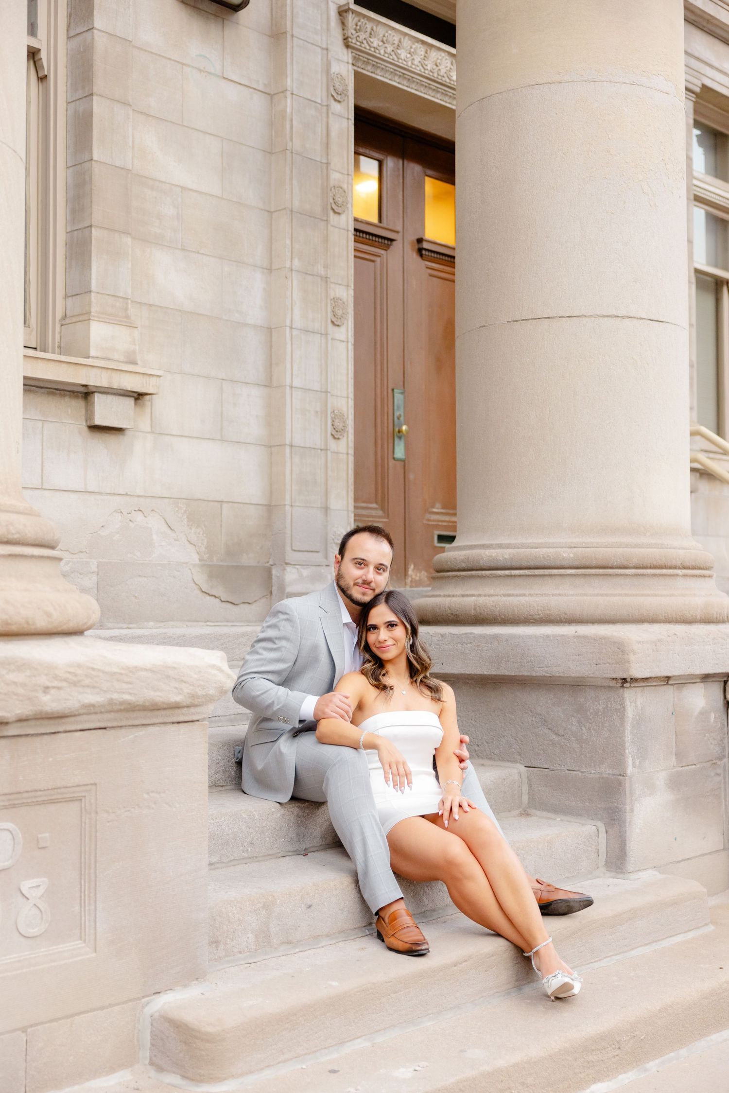 Couple relaxes on stone steps of historic building with ornate columns and wooden door.